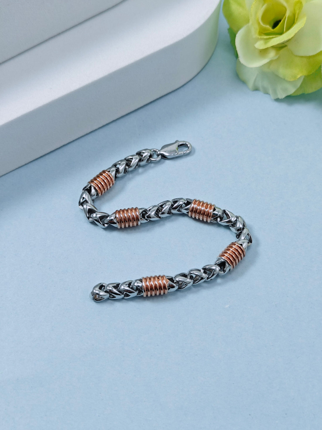 Silver and rose gold braided bracelet on a light blue surface with a white box and yellow flower in the background.