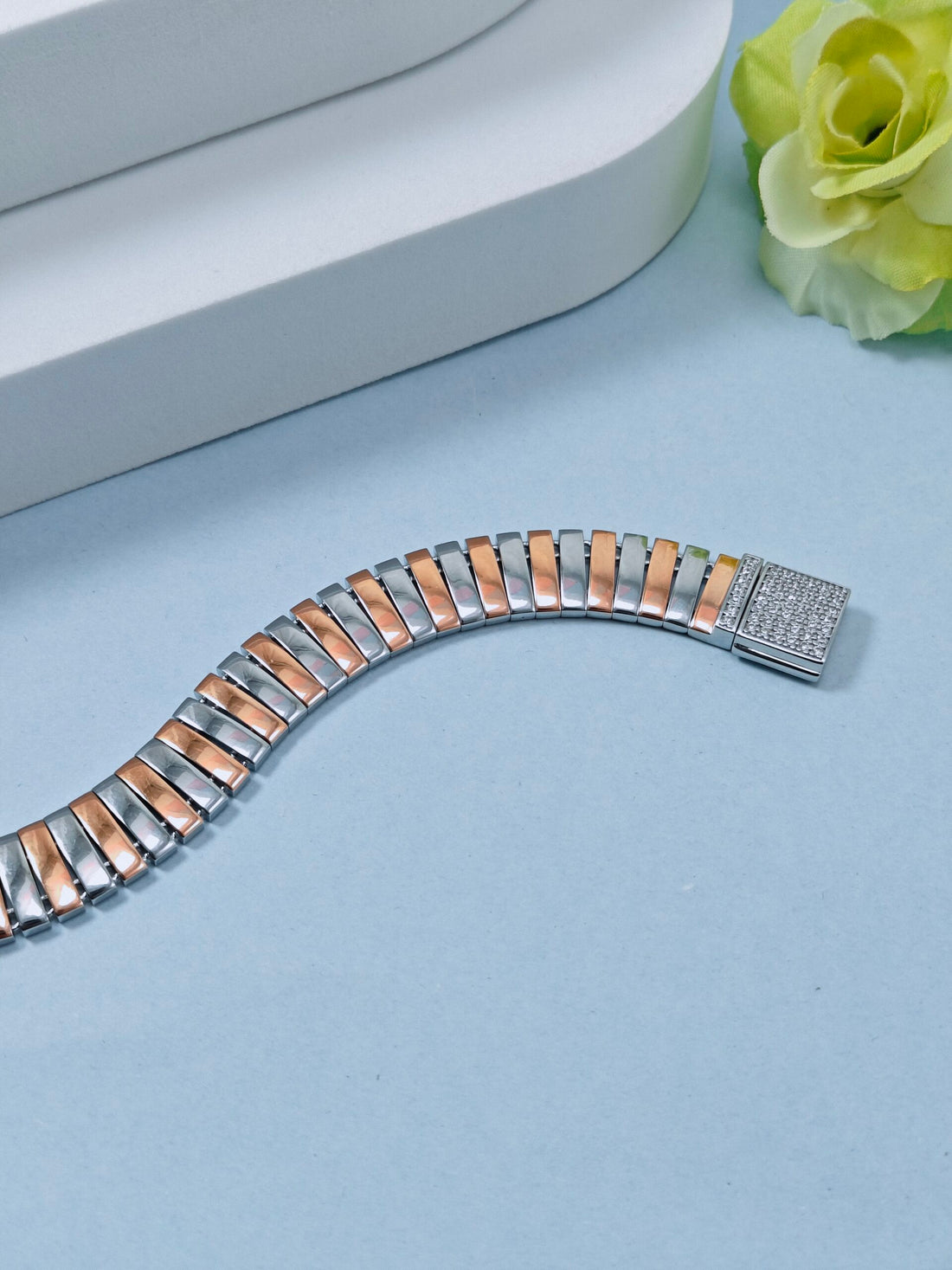 Two-tone metal bracelet on a light blue surface with a white box and green flower in the background.