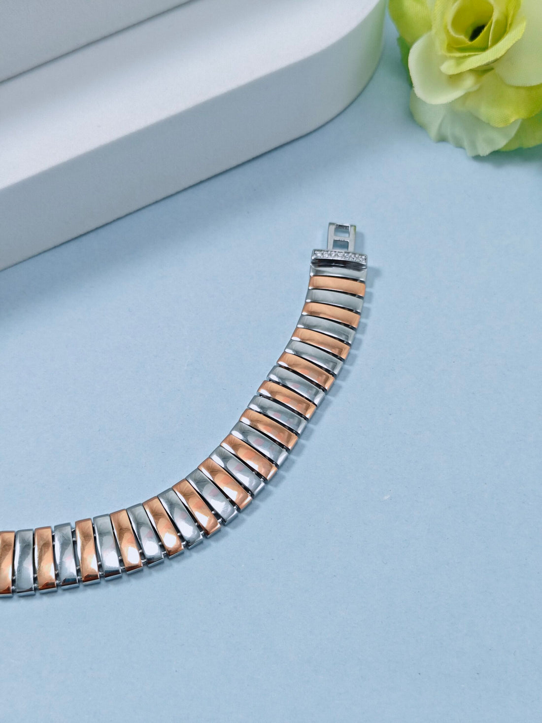 Two-tone bracelet on a light blue surface with a white box and green flower in the background