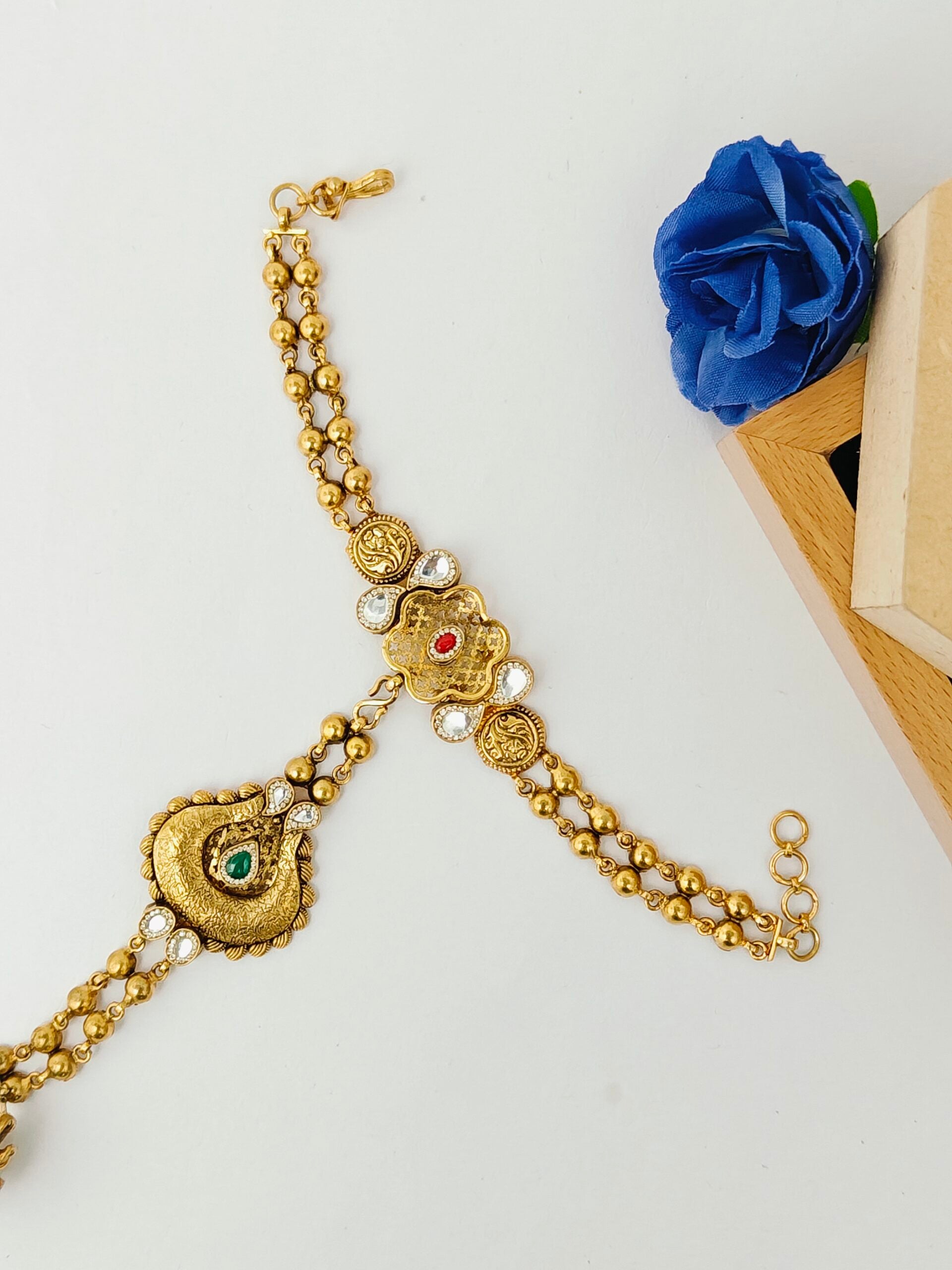 Gold bracelet with intricate designs on a white surface with a blue flower and wooden block in the background.