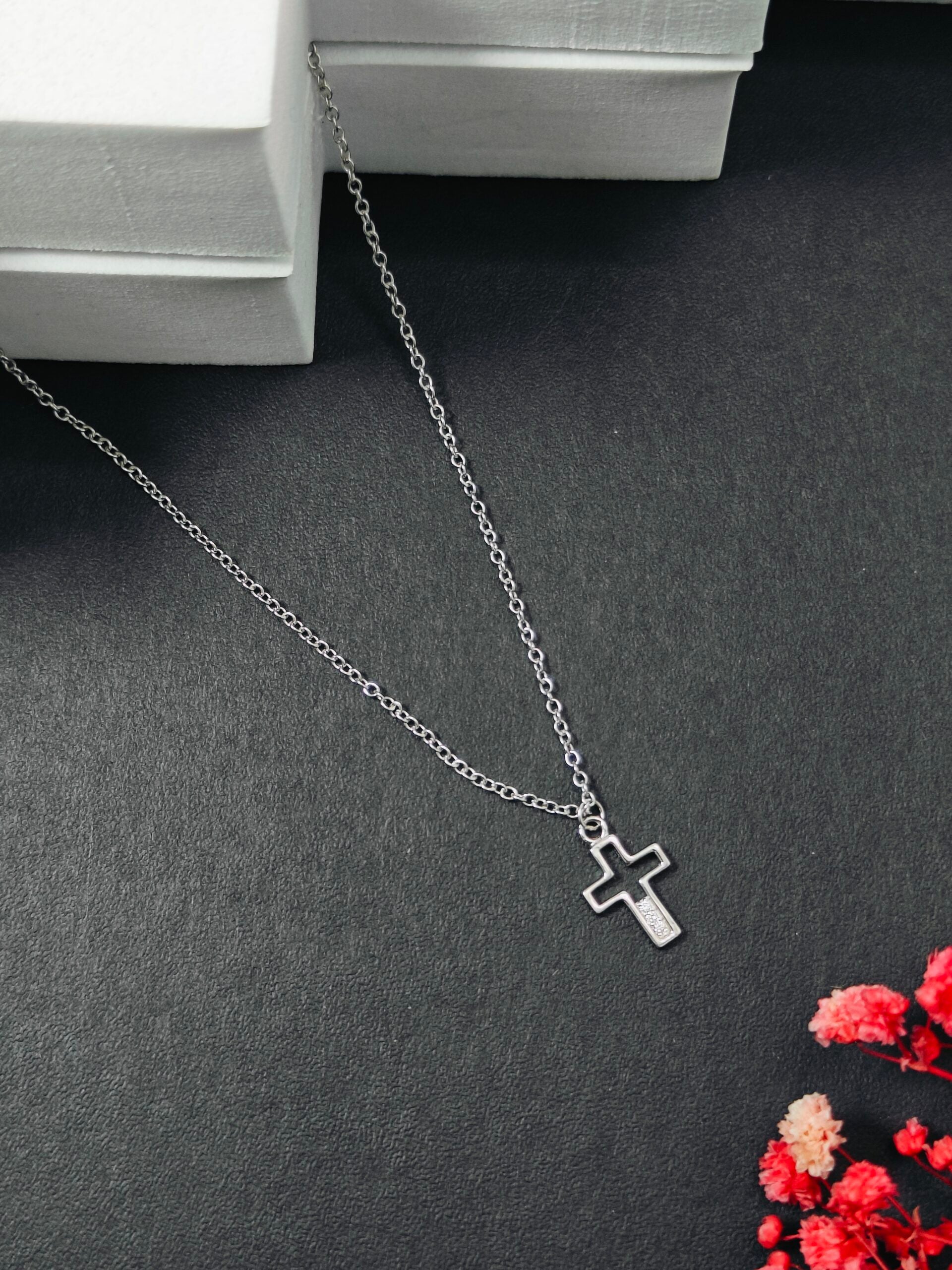 Silver necklace with a cross pendant on a dark surface with red flowers.