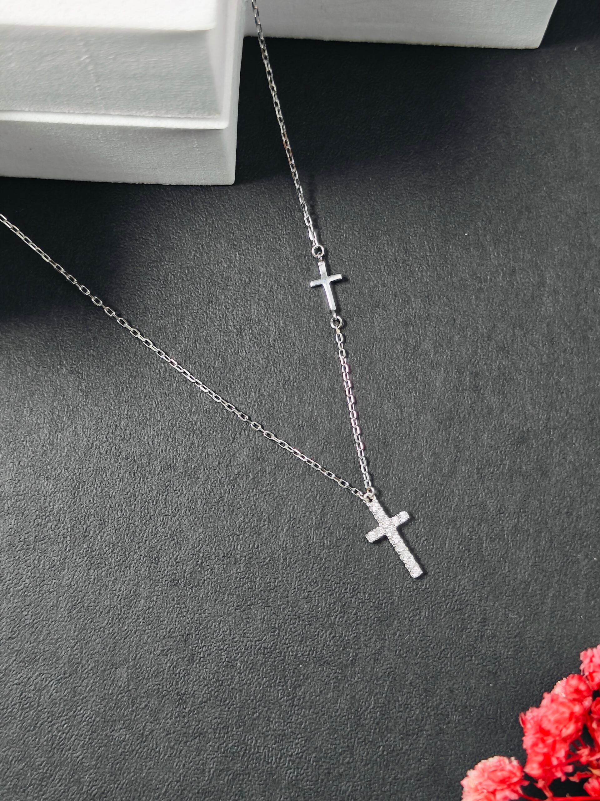 Silver cross necklace on a dark surface with a small bouquet of flowers in the corner.