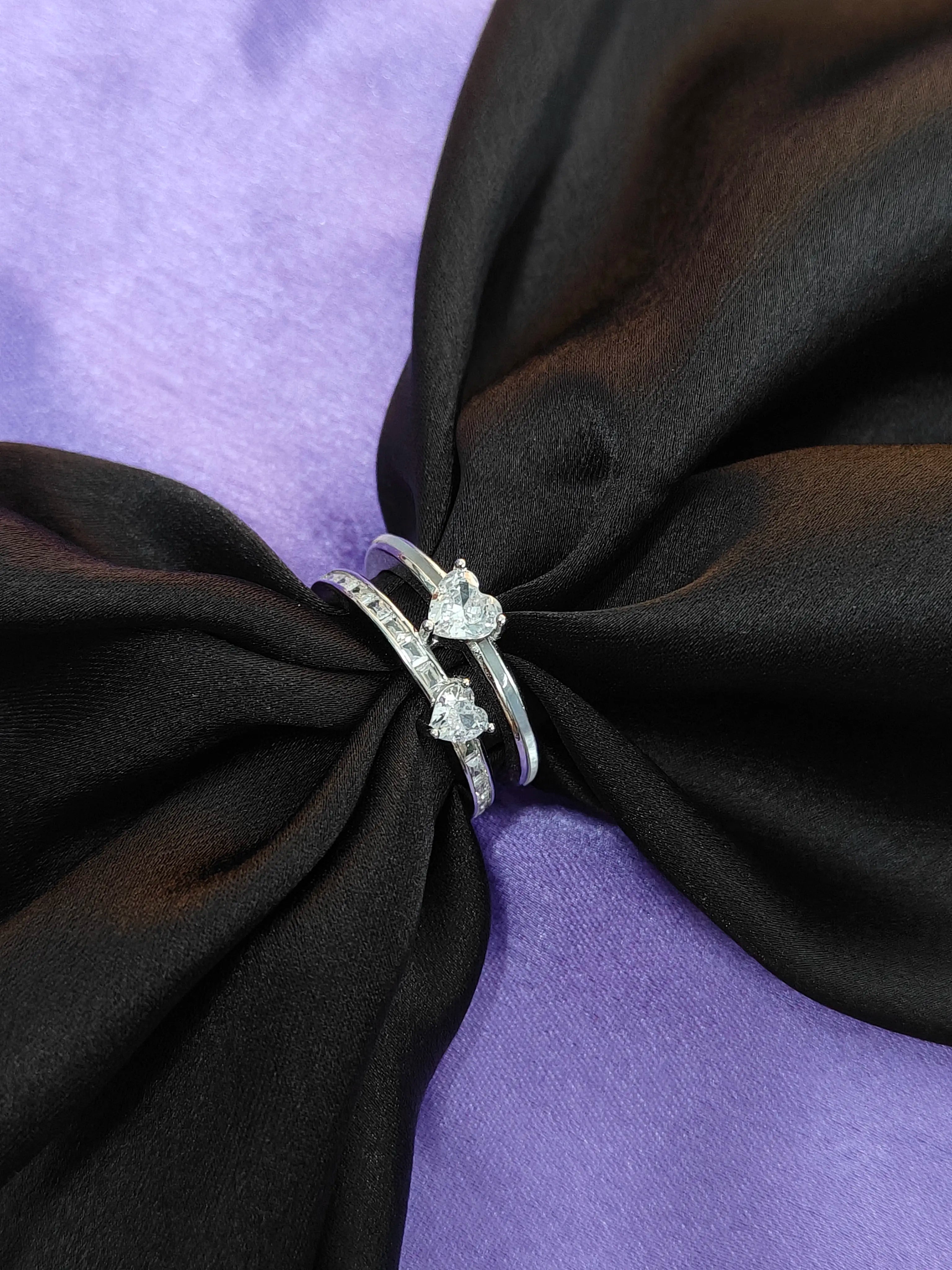 Silver ring with heart-shaped stones on a black fabric knot against a purple background