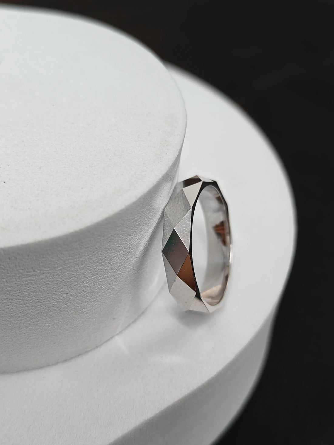 Silver ring with a faceted design on a white stand against a black background