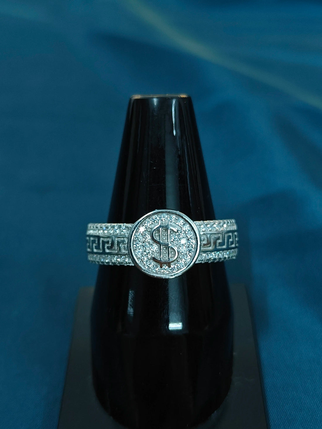Silver ring with dollar sign design on a black stand against a blue background