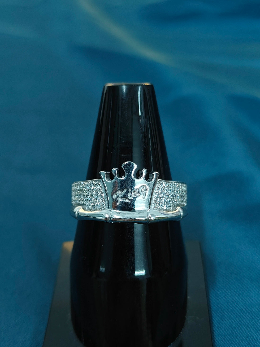 Silver ring with a crown design on a black stand against a blue background