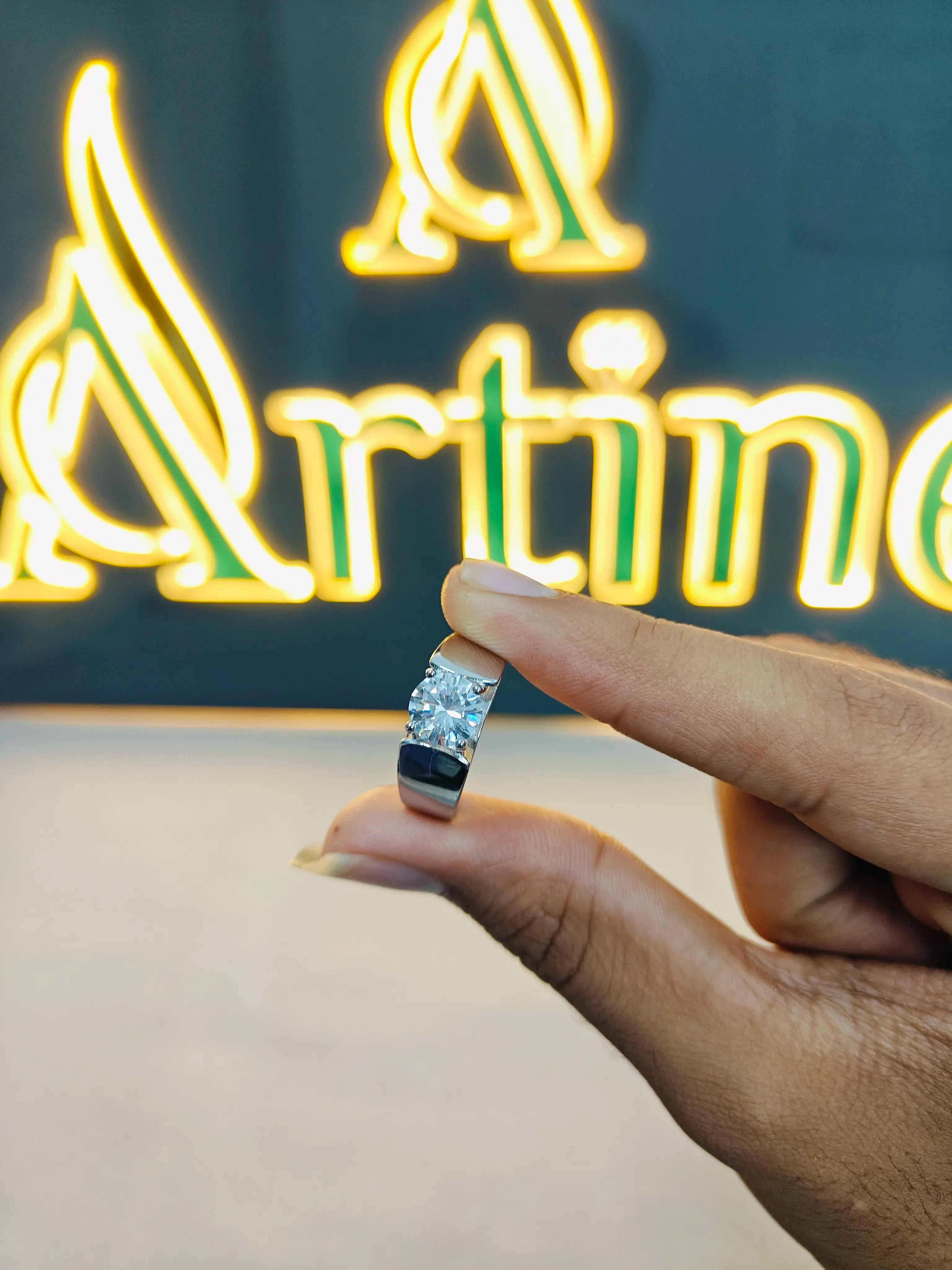 Hand holding a diamond ring in front of a neon sign with 'Artino' on a dark background