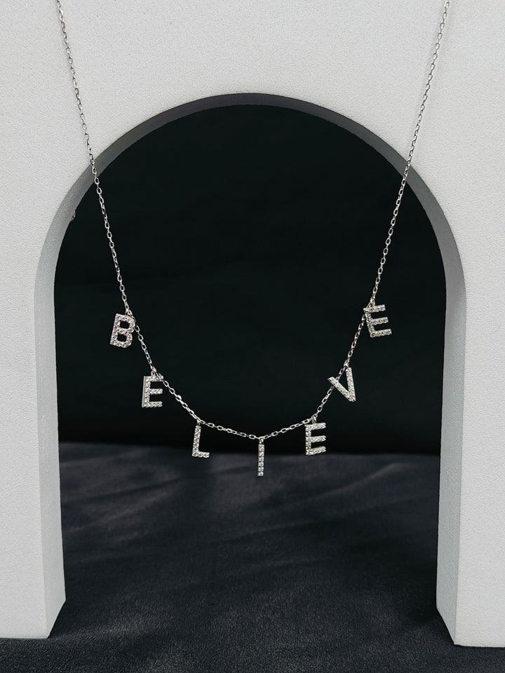 Necklace with 'BELIEVE' letters hanging in front of a black archway.