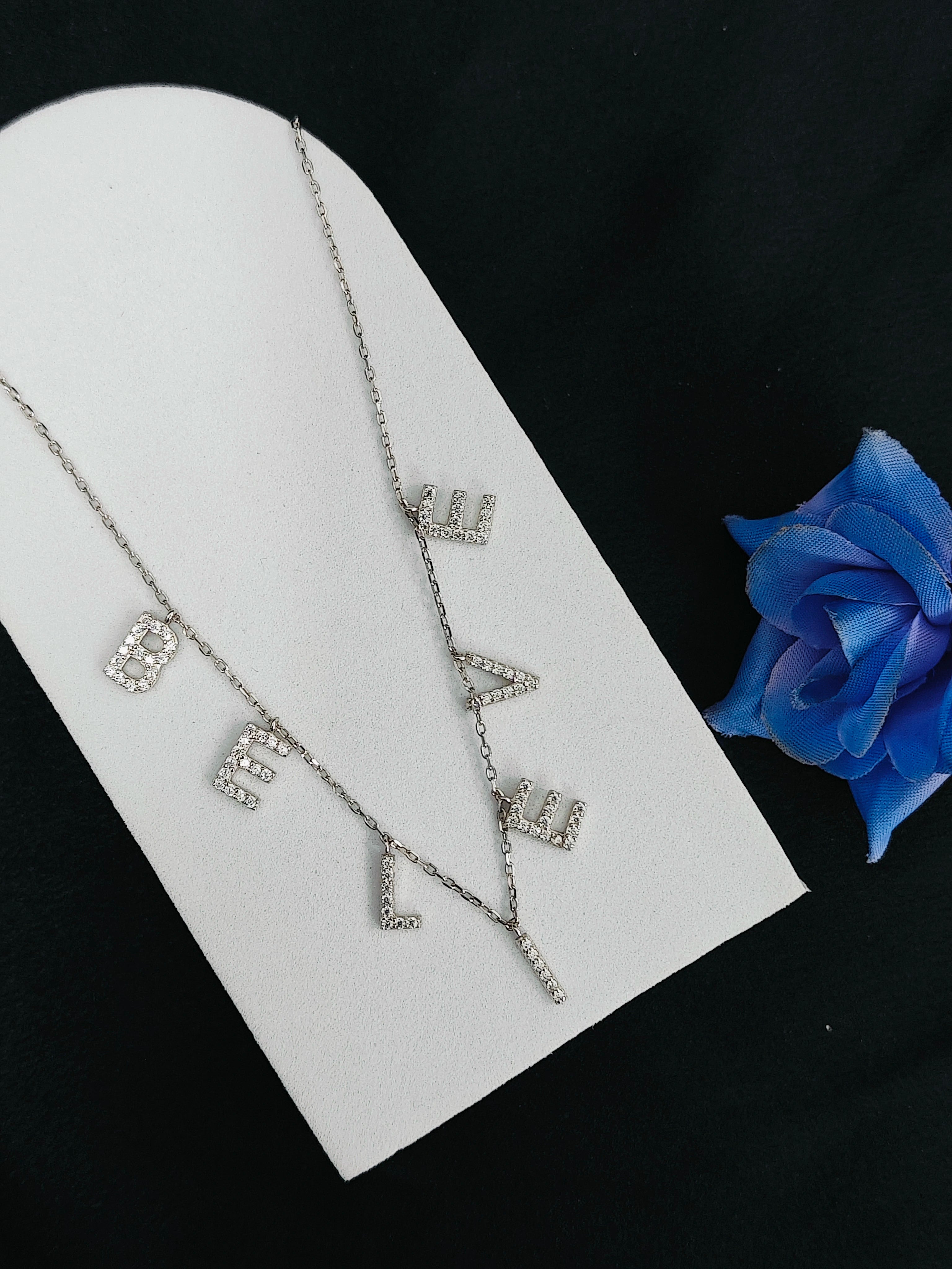 Silver necklace with letter 'V' design on a white stand with a blue rose.