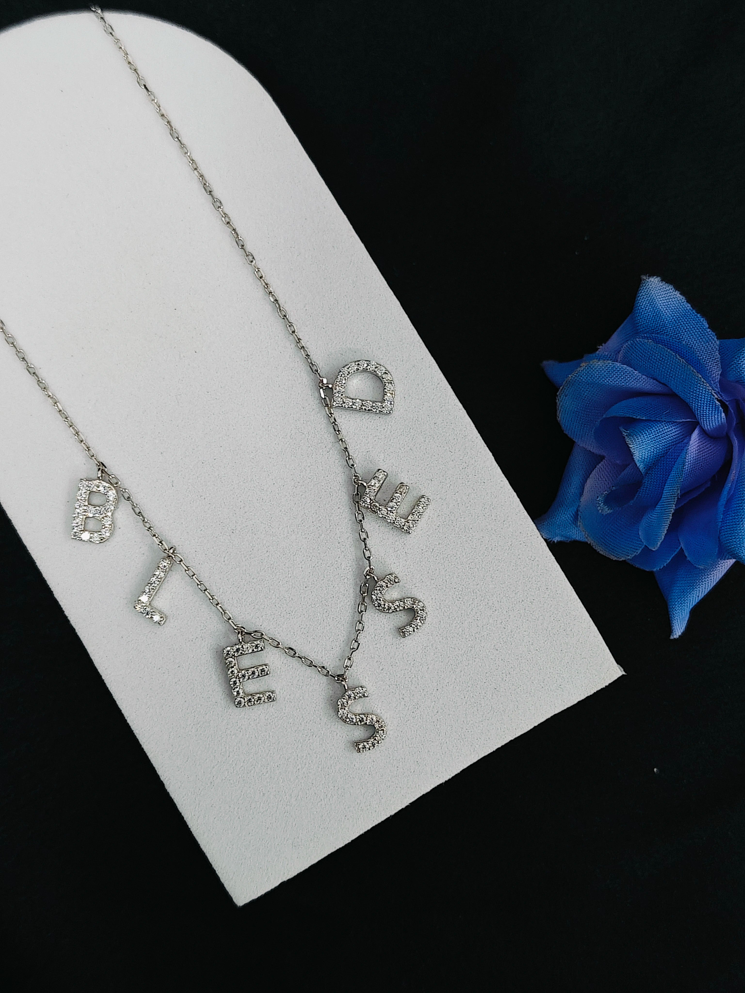 Necklace with letter charms on a white card with a blue rose in the background