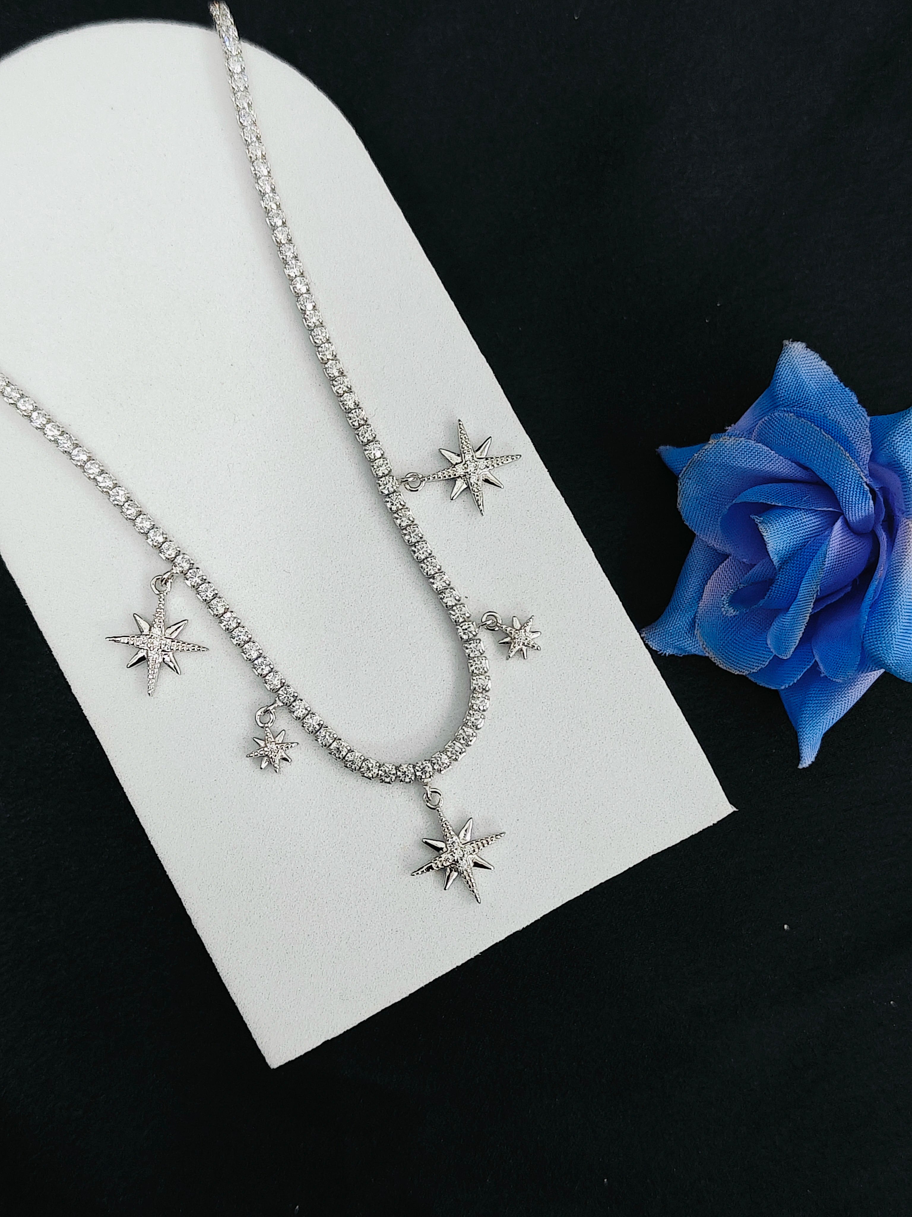 Silver necklace with star-shaped pendants on a white stand with a blue flower.