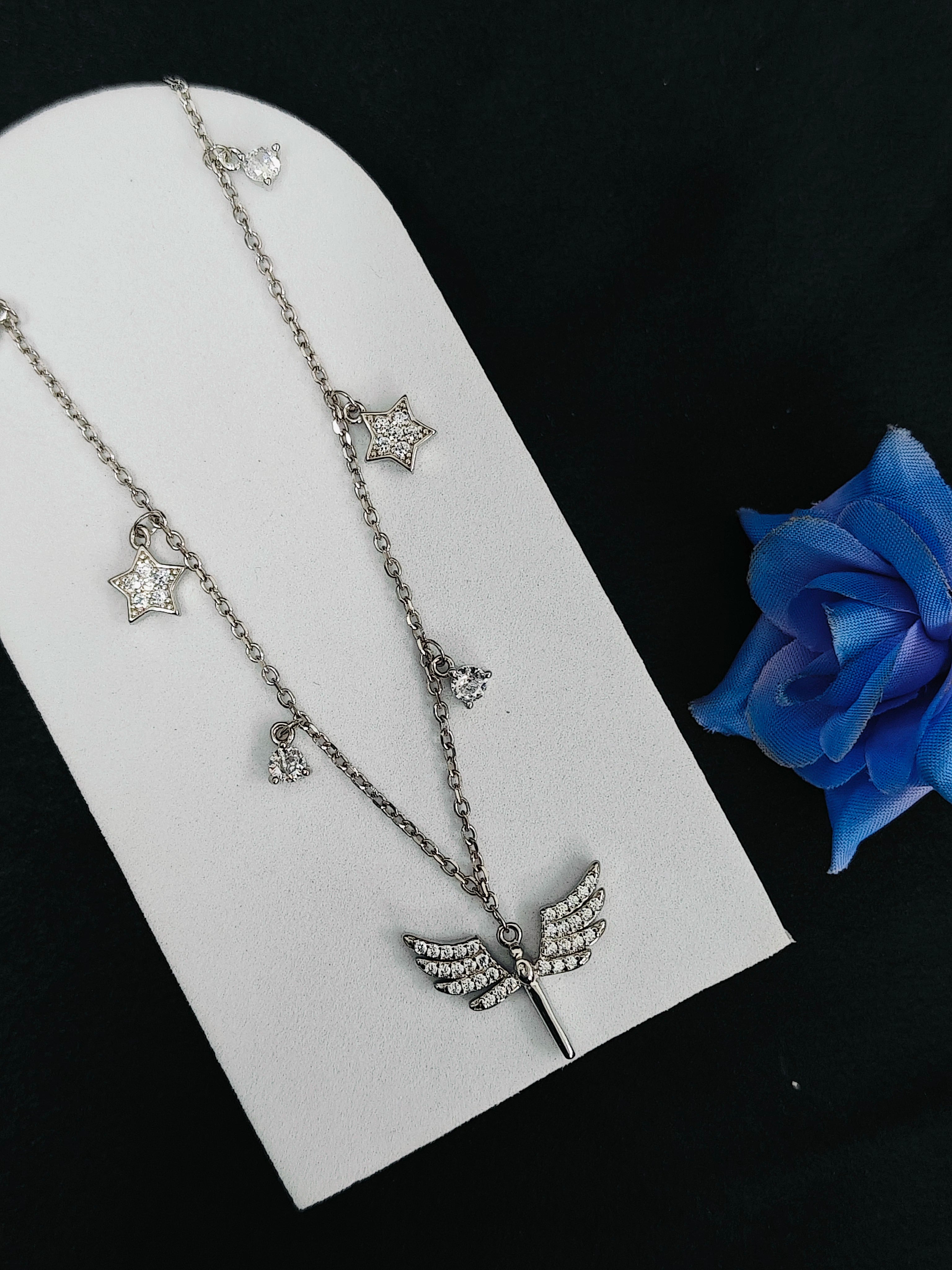 Silver necklace with star and leaf pendants on a white stand with a blue rose.