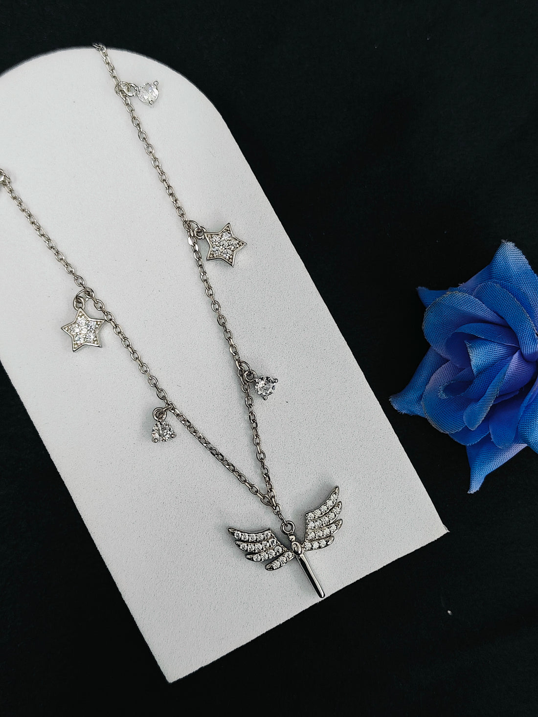 Silver necklace with star and leaf pendants on a white stand with a blue rose.