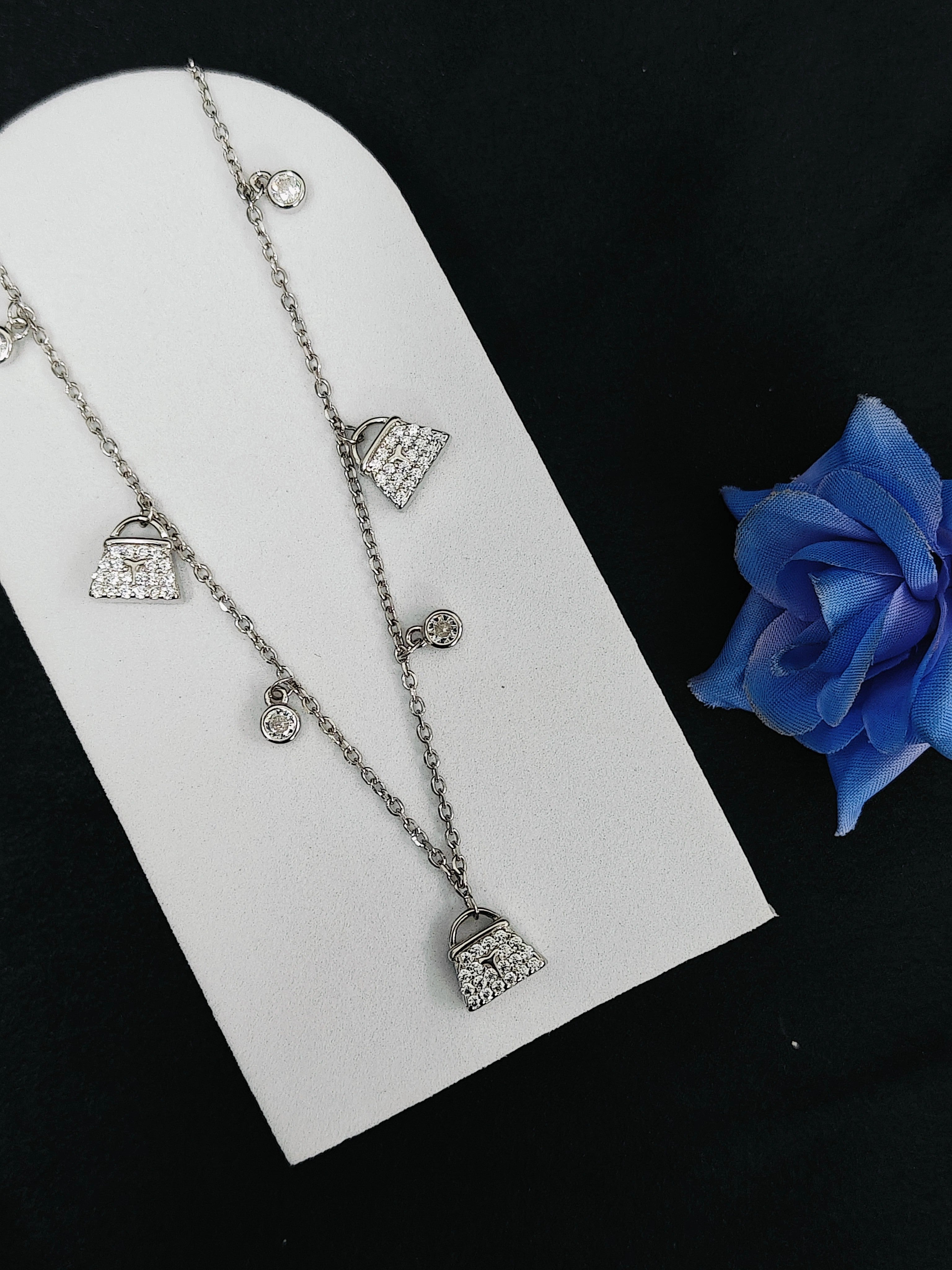 Silver necklace with heart and bag charms on a white stand with a blue rose.