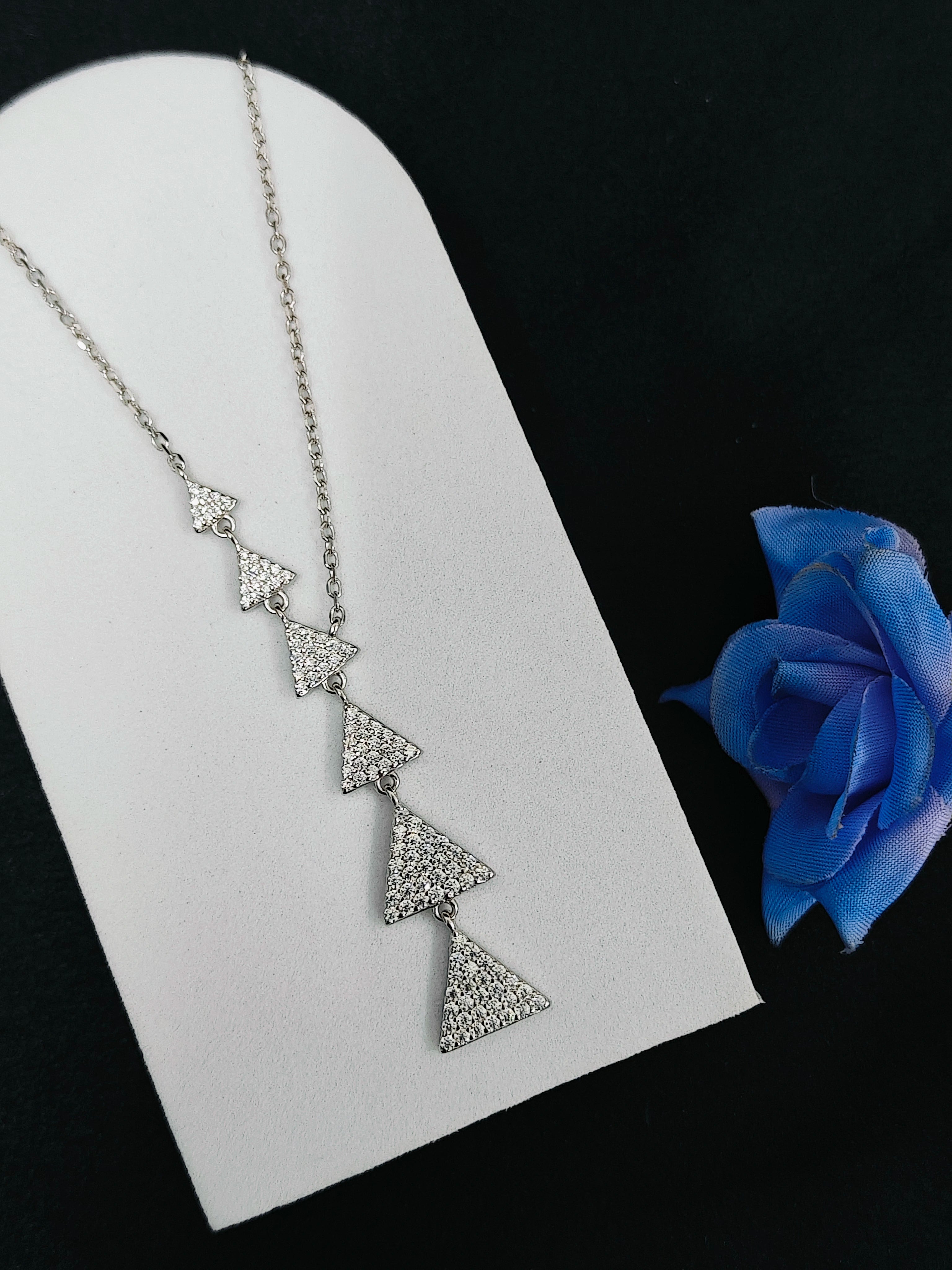 Silver necklace with triangular pendants on a white stand with a blue rose.
