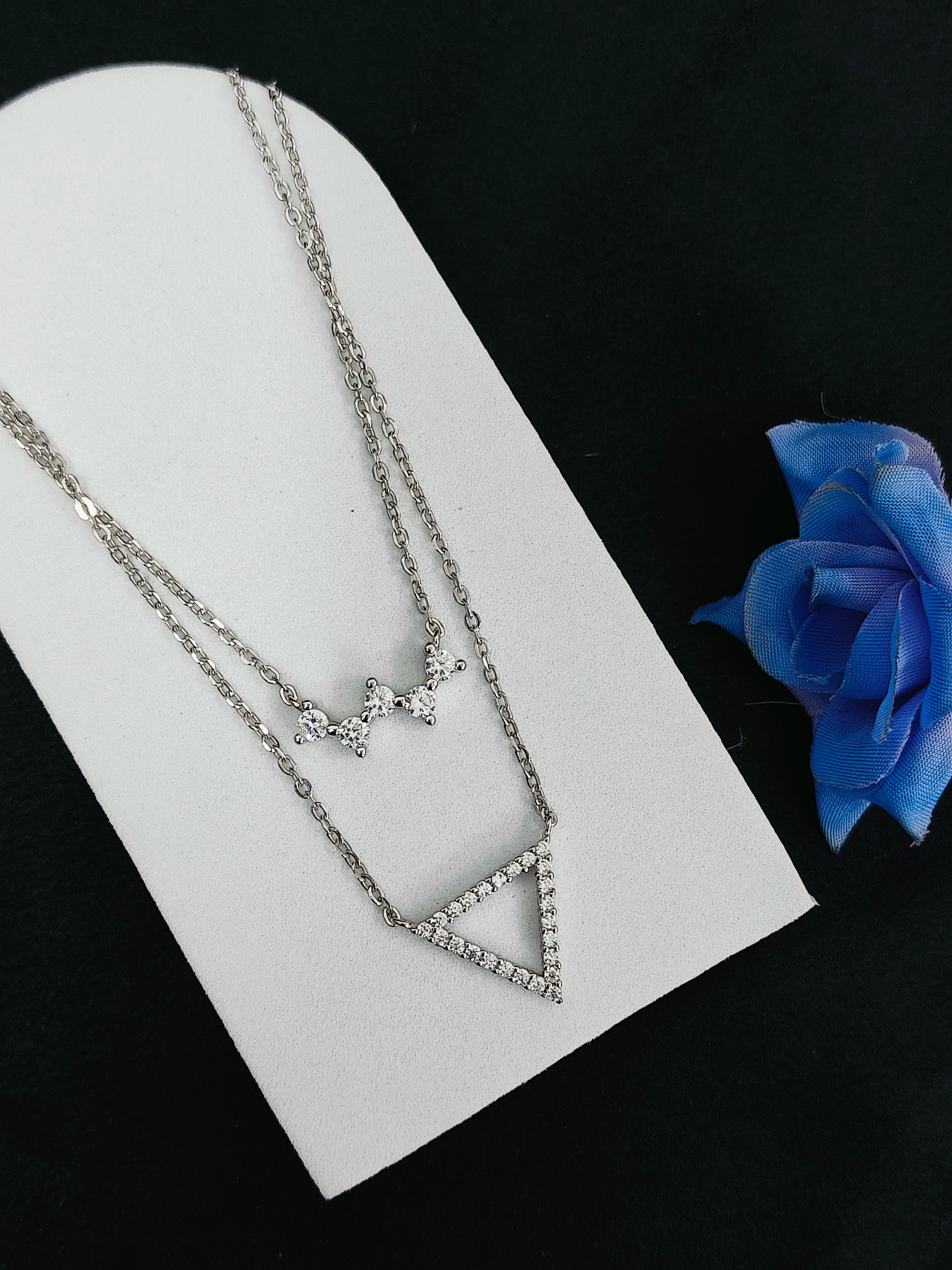Silver necklace with star and triangle design on a white stand with a blue rose.