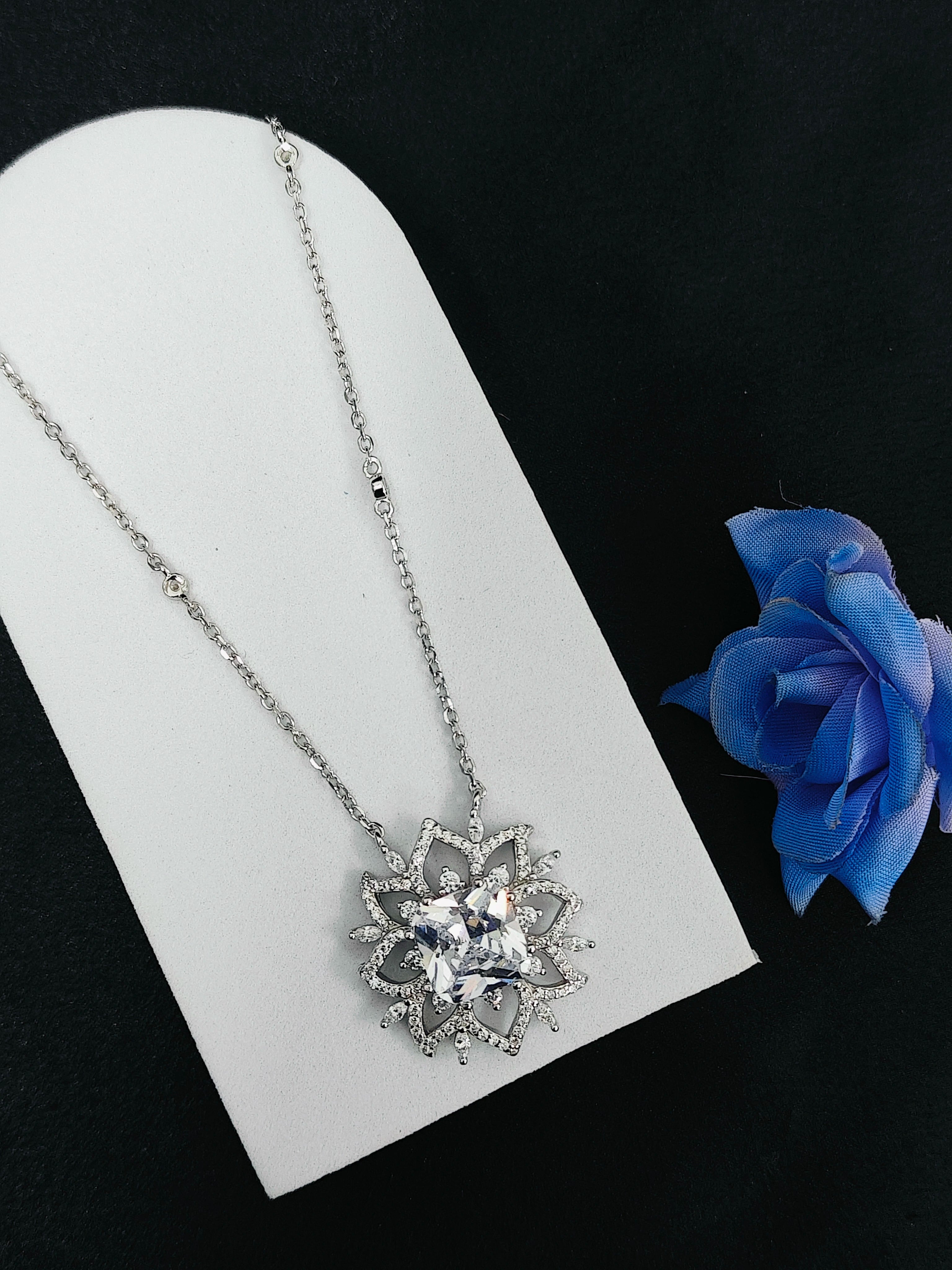 Silver necklace with a star-shaped pendant on a white stand next to a blue rose.
