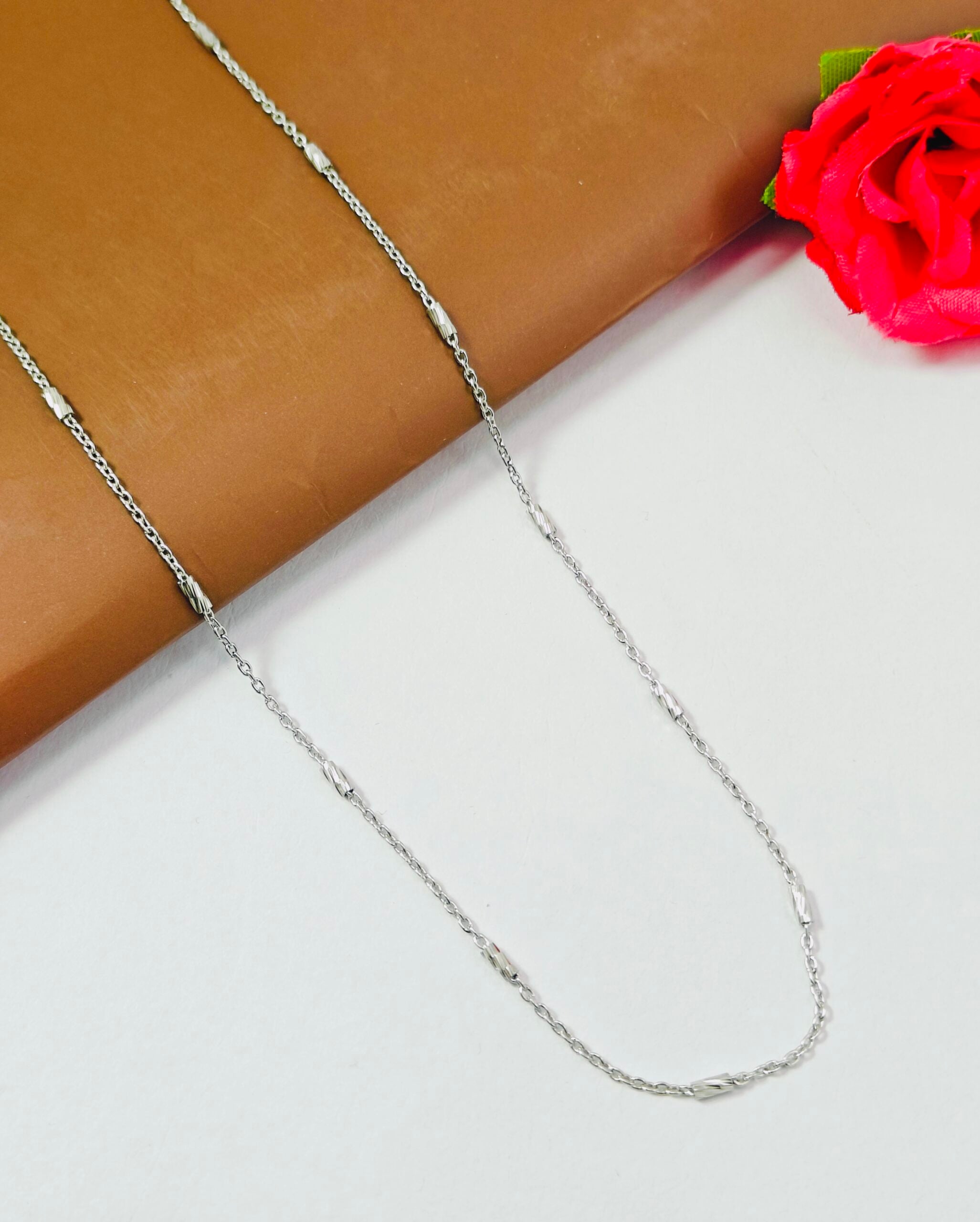 Silver chain necklace on a white surface with a red flower in the corner