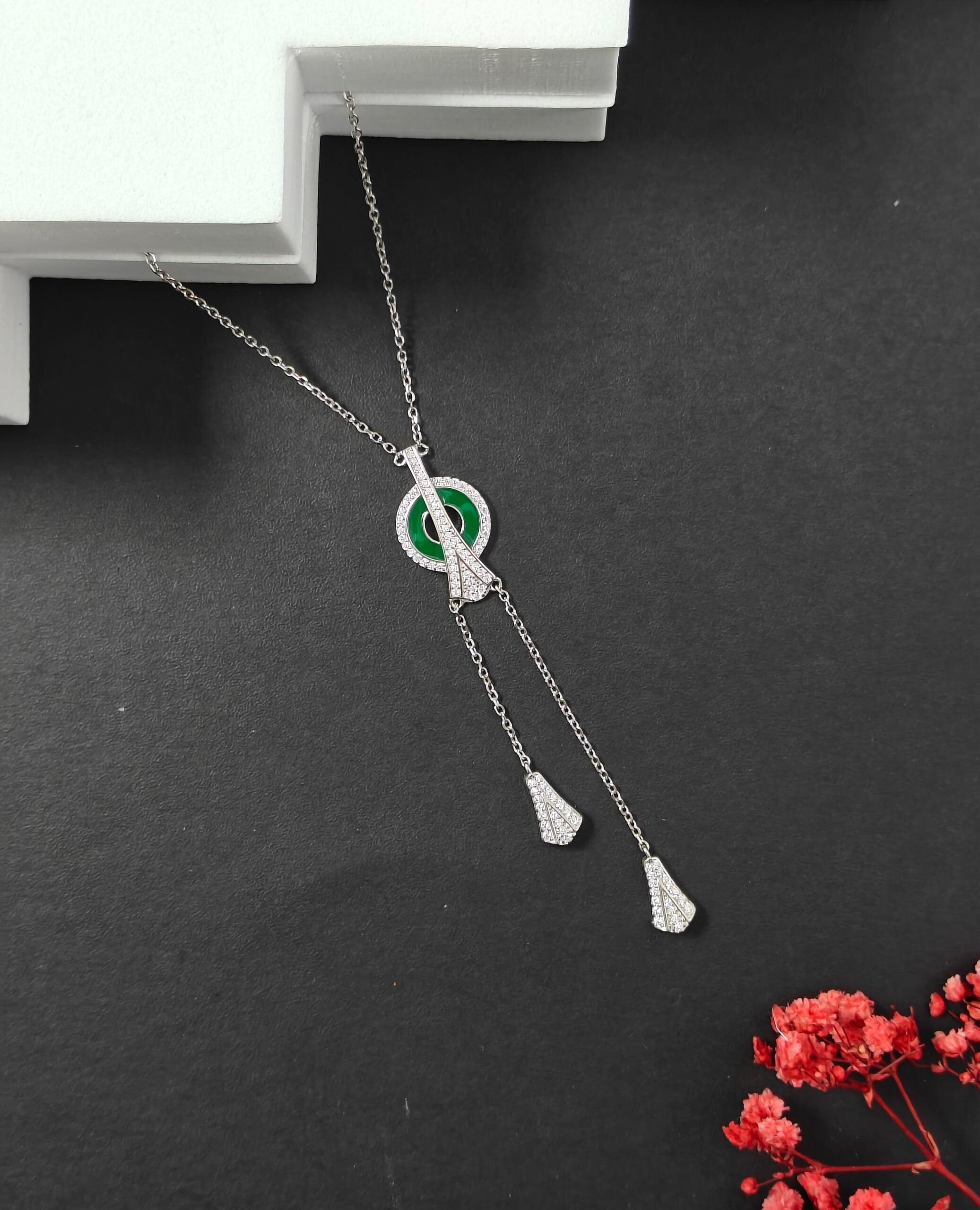 Silver necklace with green gemstone on a dark surface with red flowers.