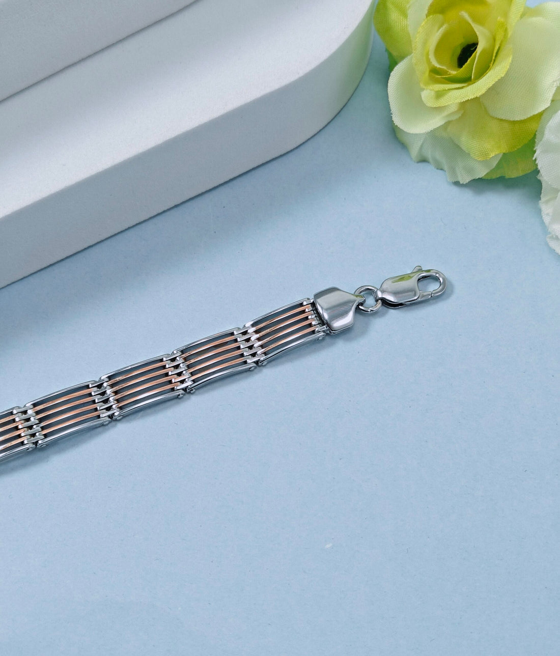 Silver bracelet with a heart clasp on a light blue surface with a white box and yellow flowers in the background.