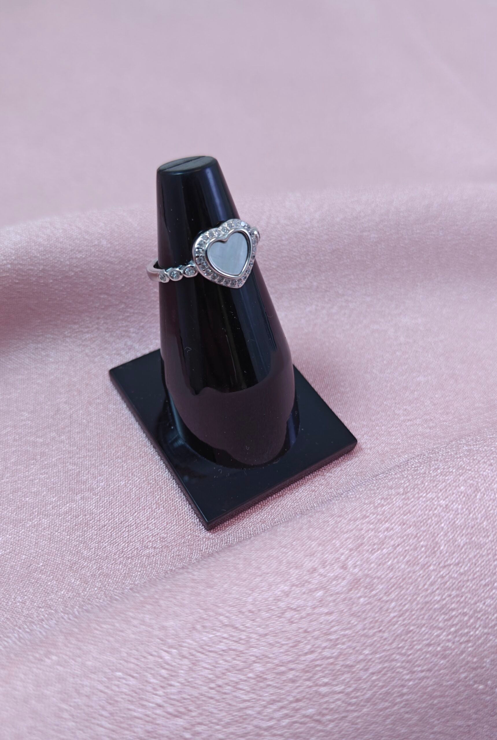 Silver ring with a heart-shaped stone on a black stand against a pink fabric background