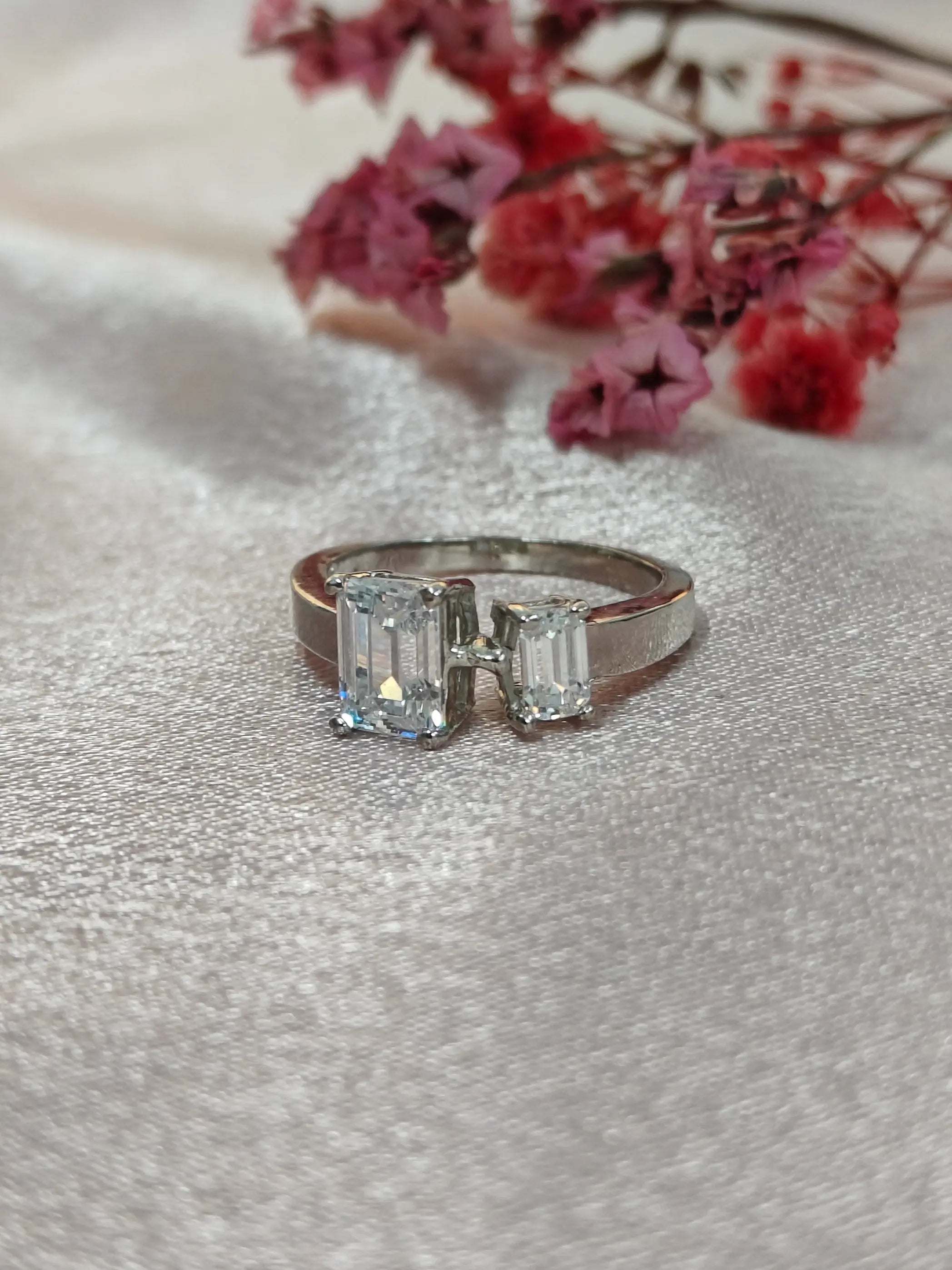 Diamond ring with emerald-cut stones on a textured surface with pink flowers in the background