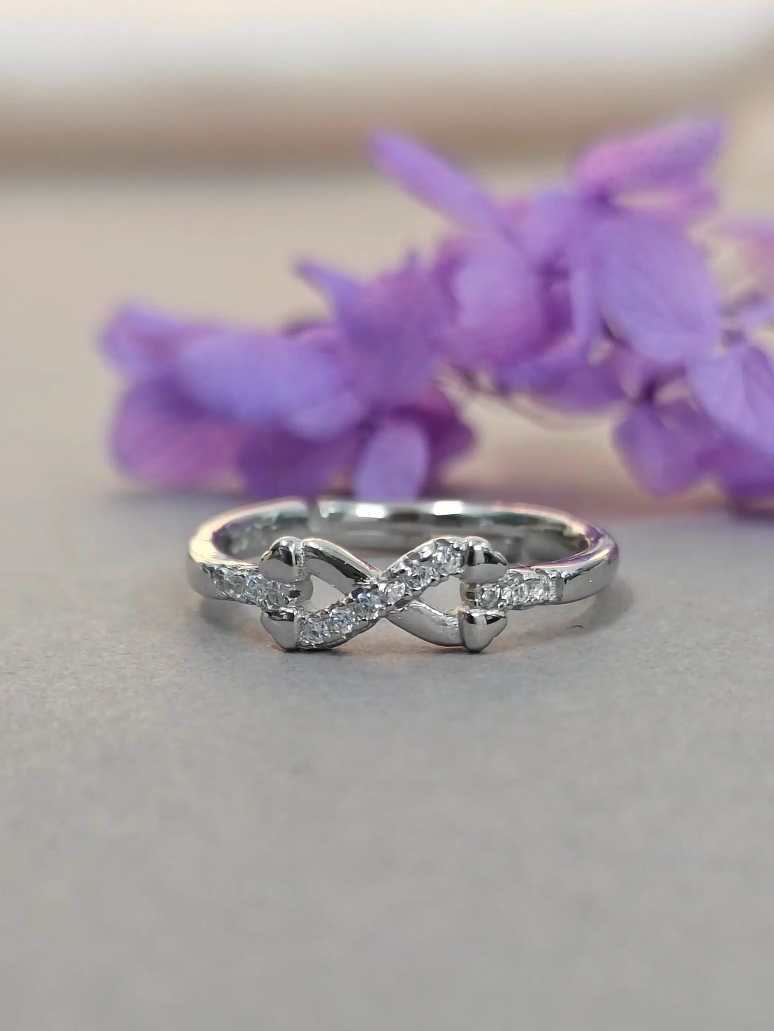 Silver infinity ring with diamonds on a gray surface with purple flowers in the background