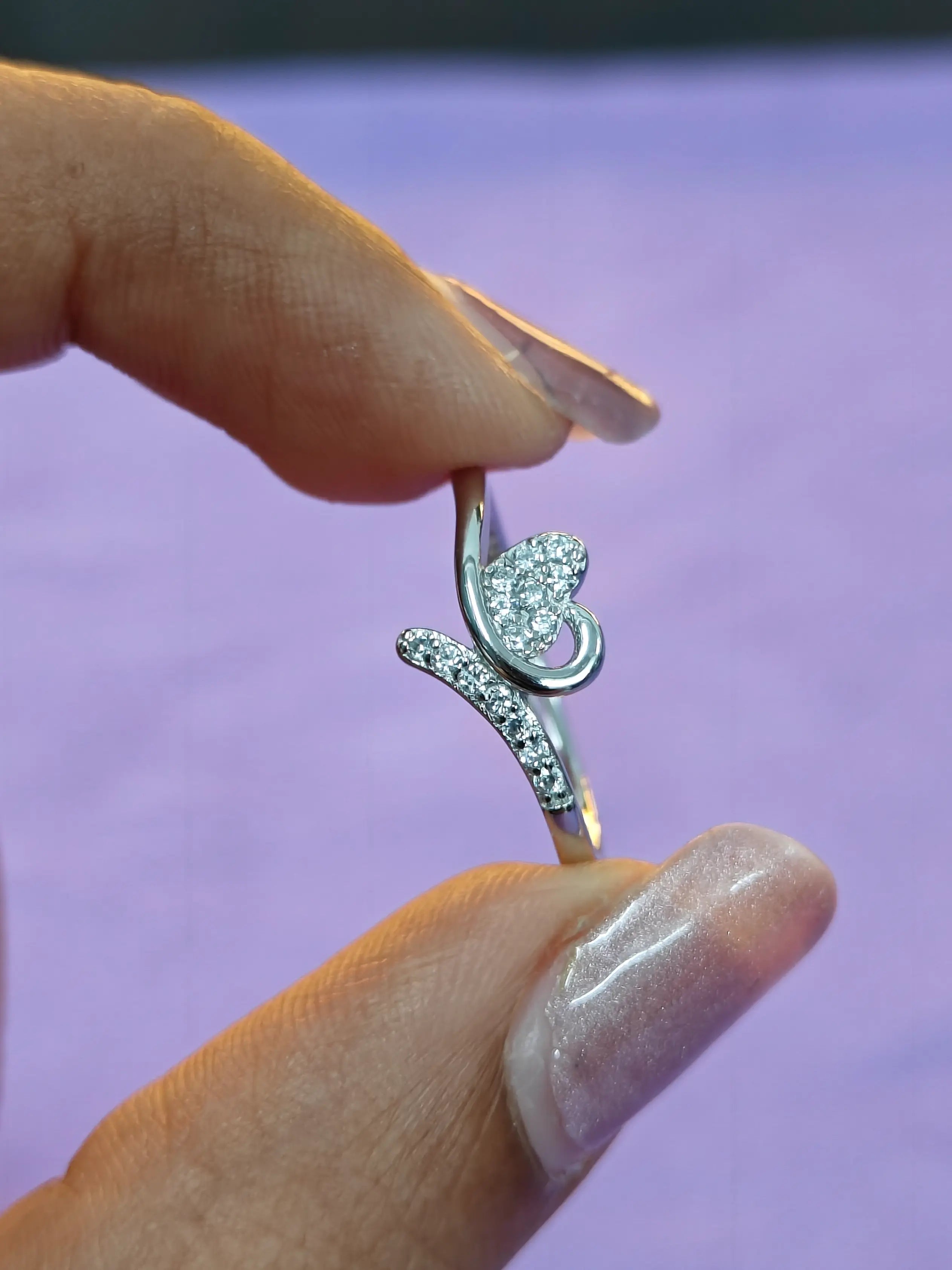 Silver ring with a heart-shaped diamond held by a hand against a purple background