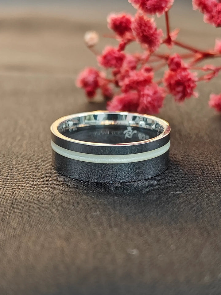 Black ring with silver and green accents on a textured surface with pink flowers in the background