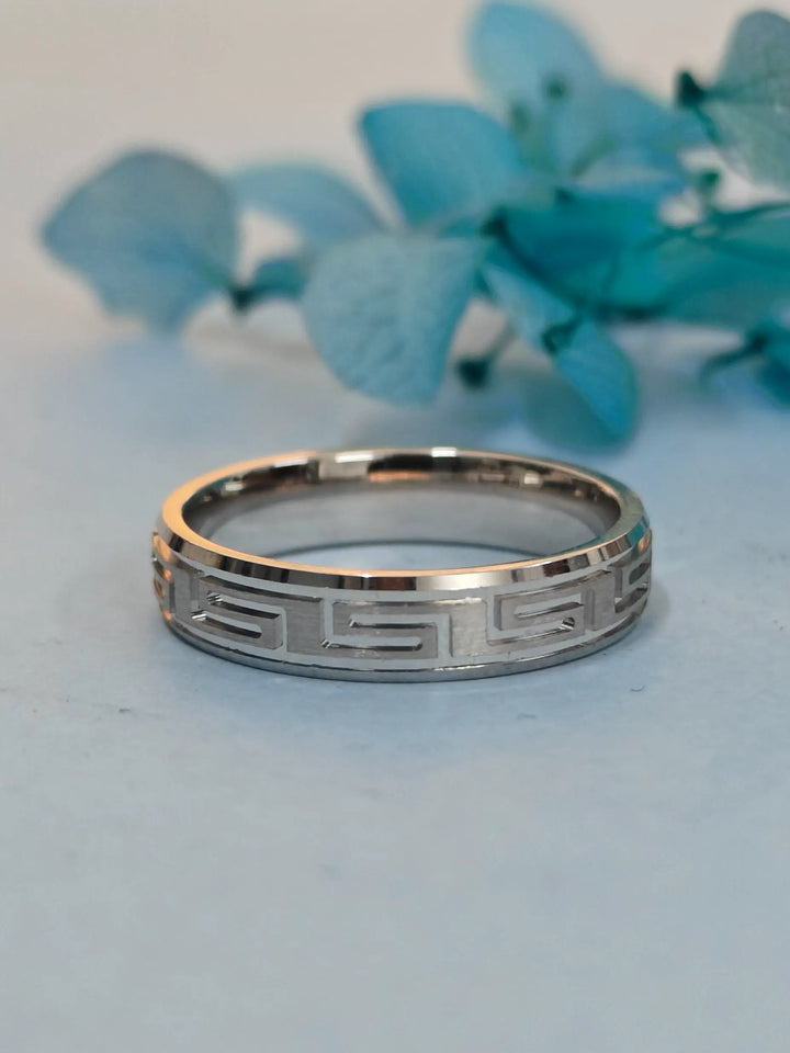 Silver ring with Greek key design on a light blue background with teal flowers.