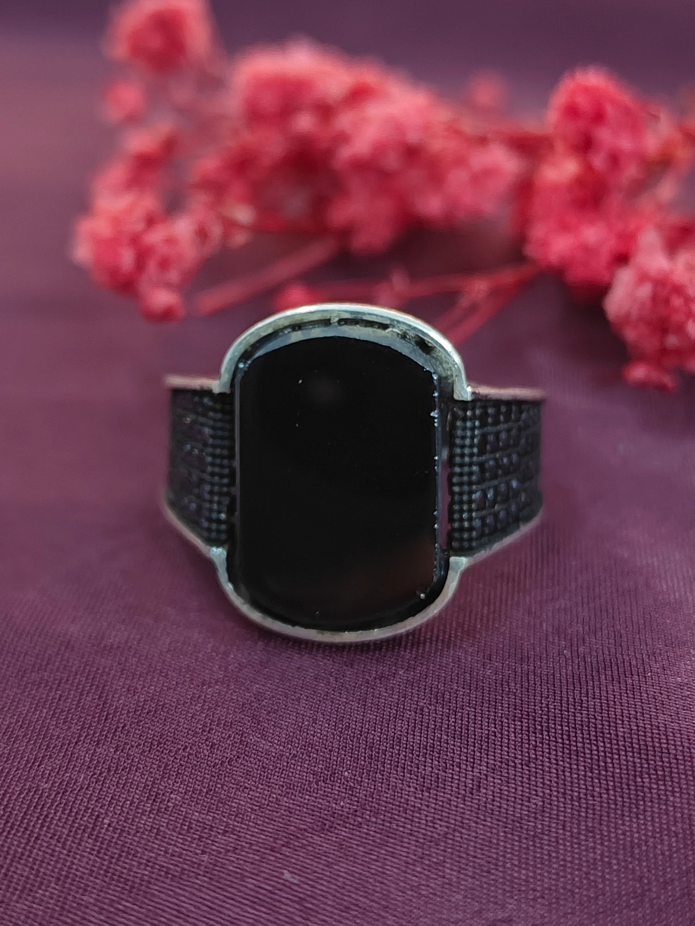 Black rectangular ring with textured band on a purple fabric background with pink flowers.