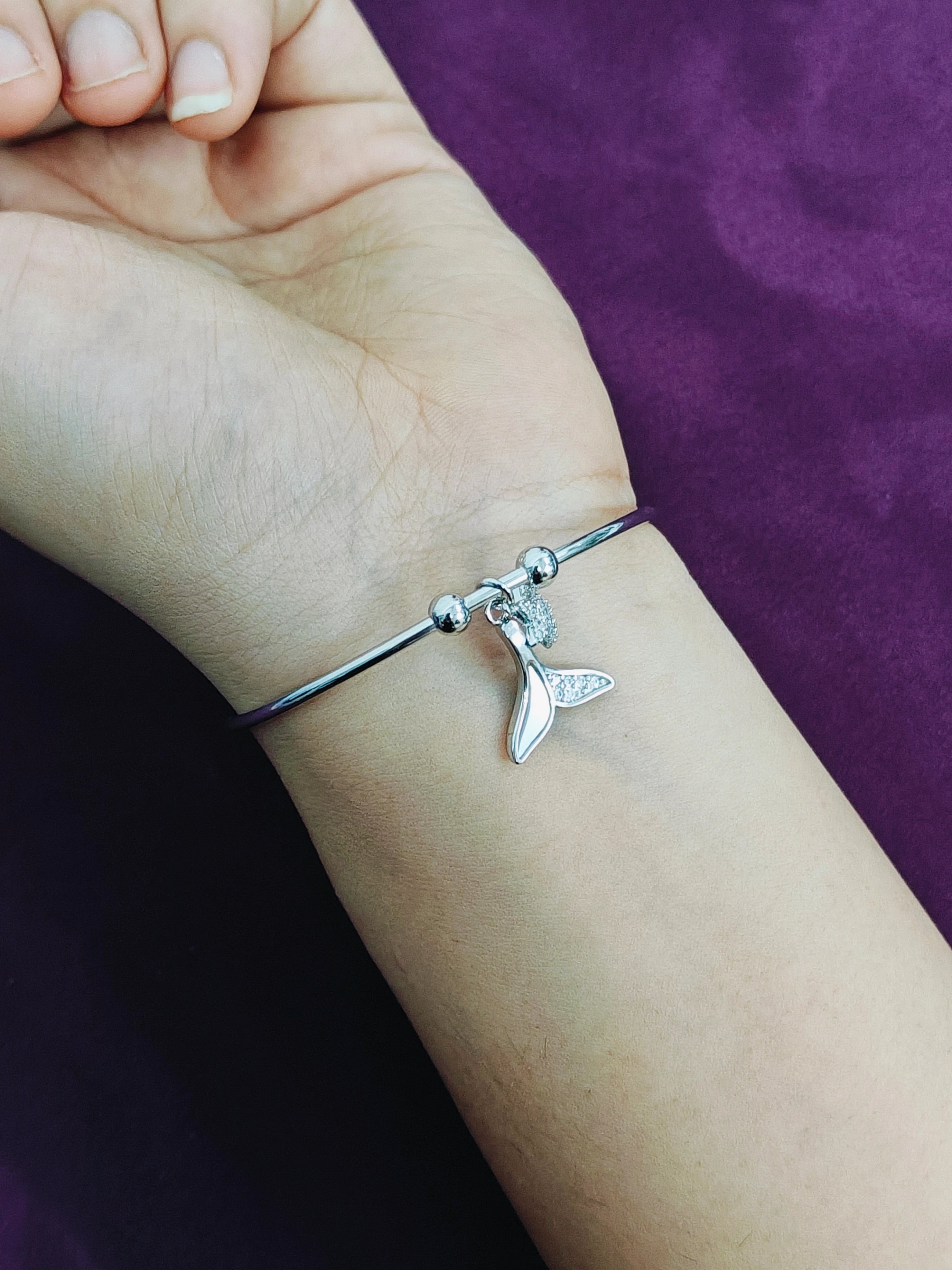 Silver bracelet with a whale tail charm on a wrist against a purple background