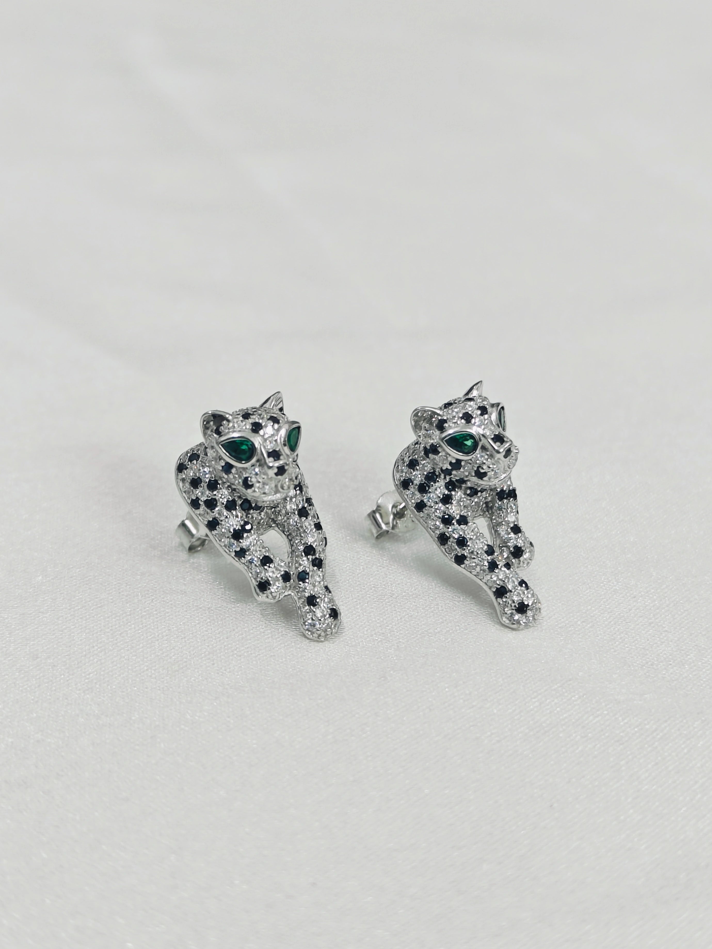 Pair of silver panther-shaped earrings with green and black gemstones on a light gray background