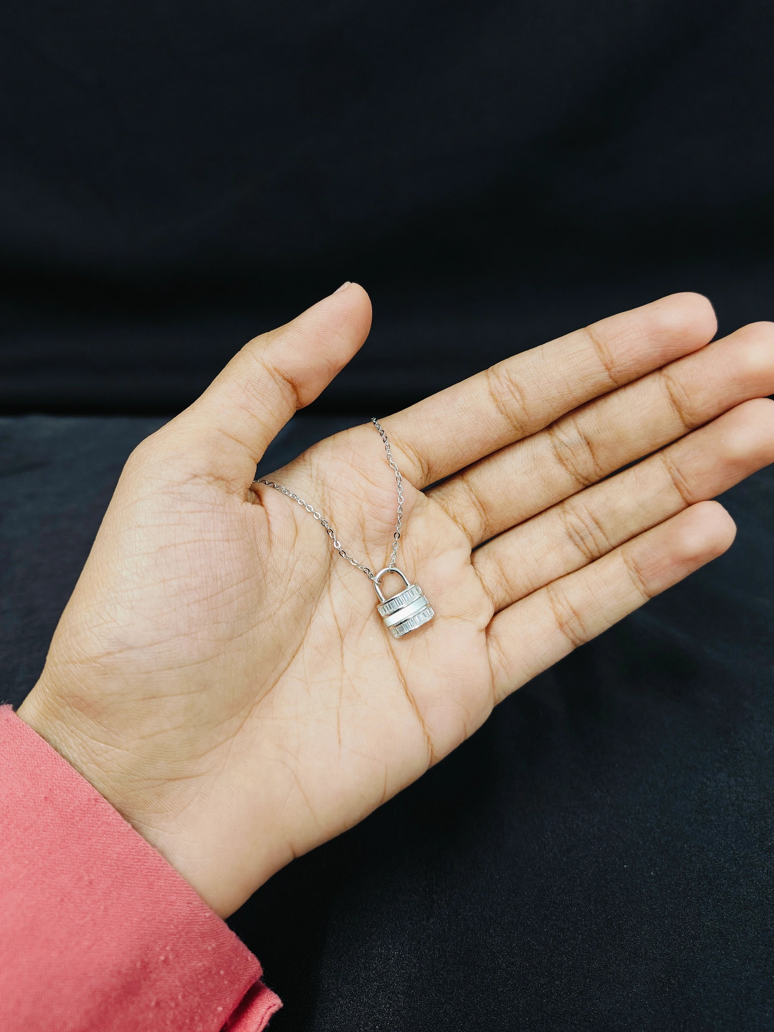 Hand holding a delicate silver necklace with a lock pendant against a black background