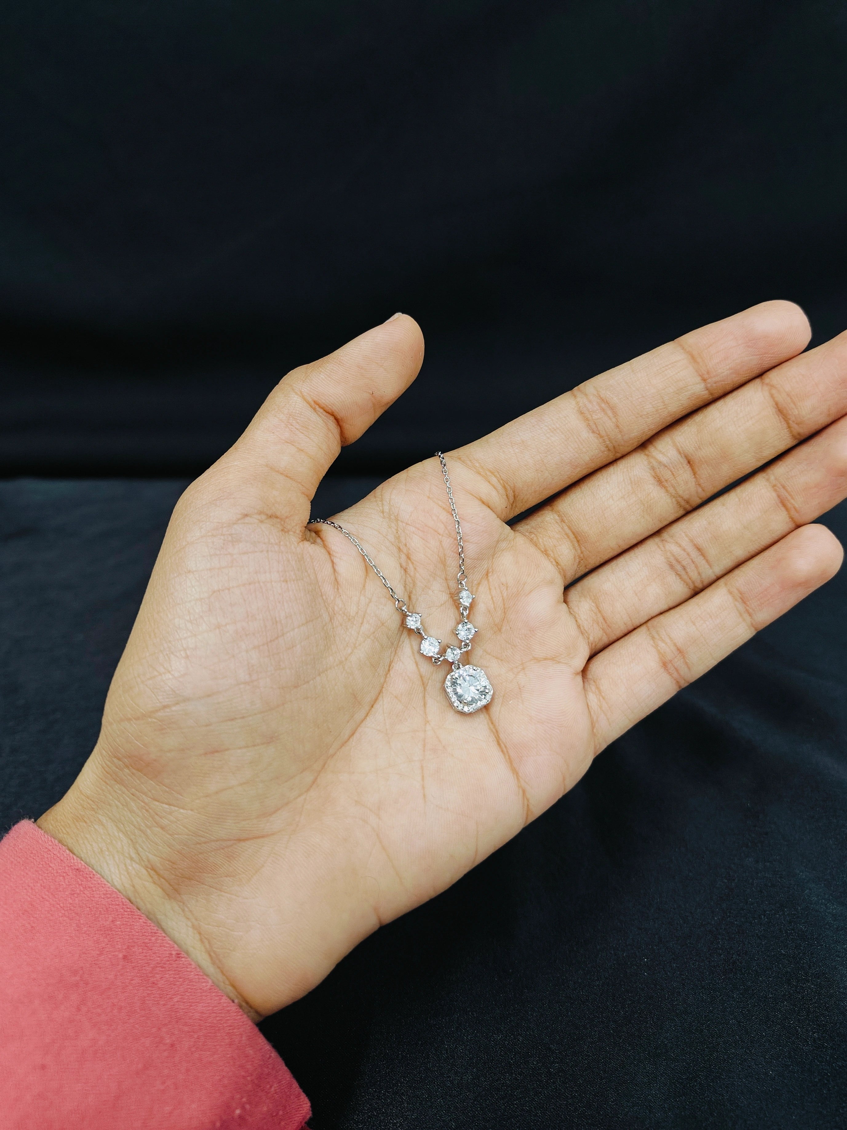 Hand holding a delicate silver necklace with a clear pendant against a dark background