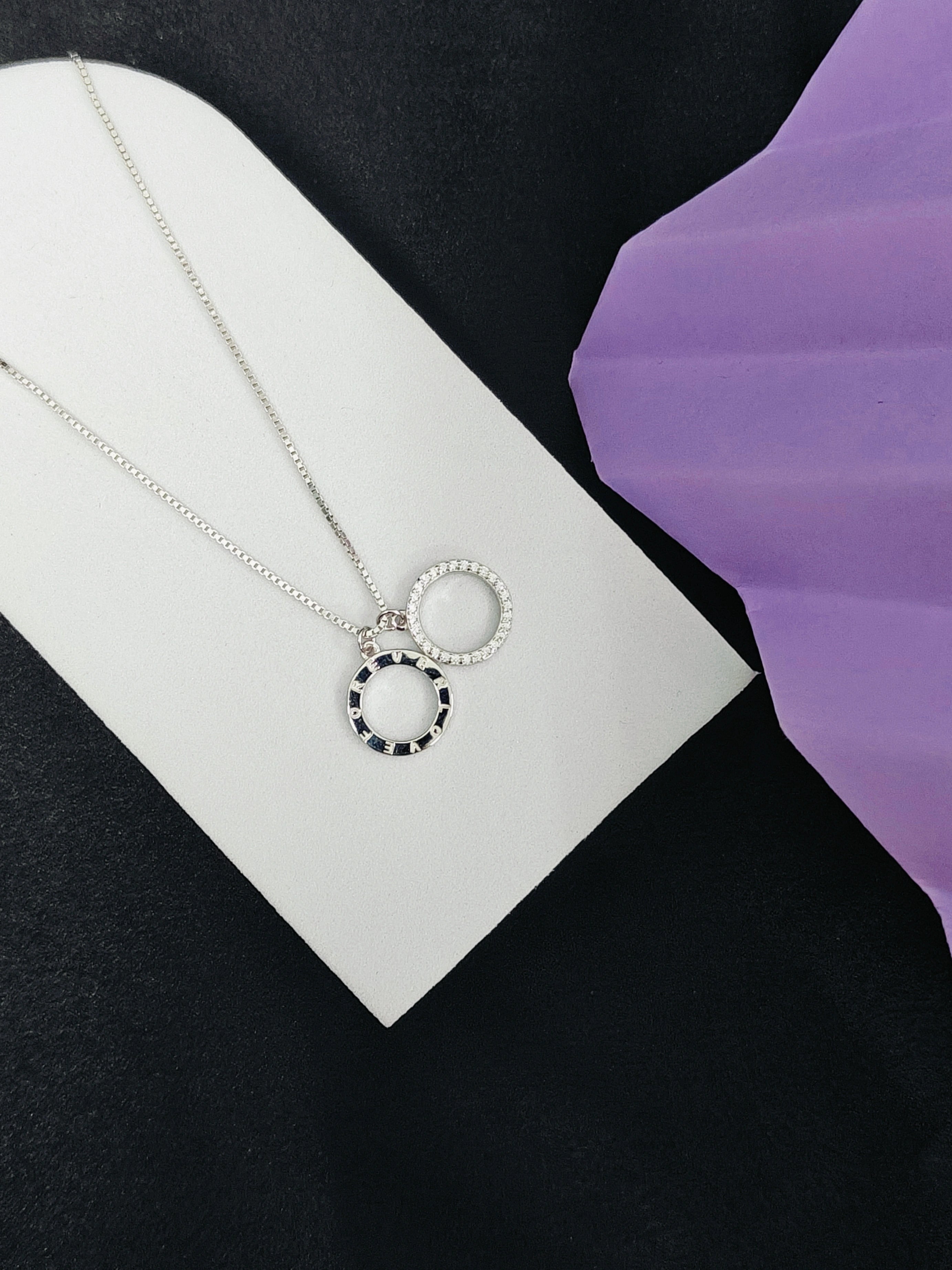 Silver necklace with a circular pendant on a white card against a black and purple background