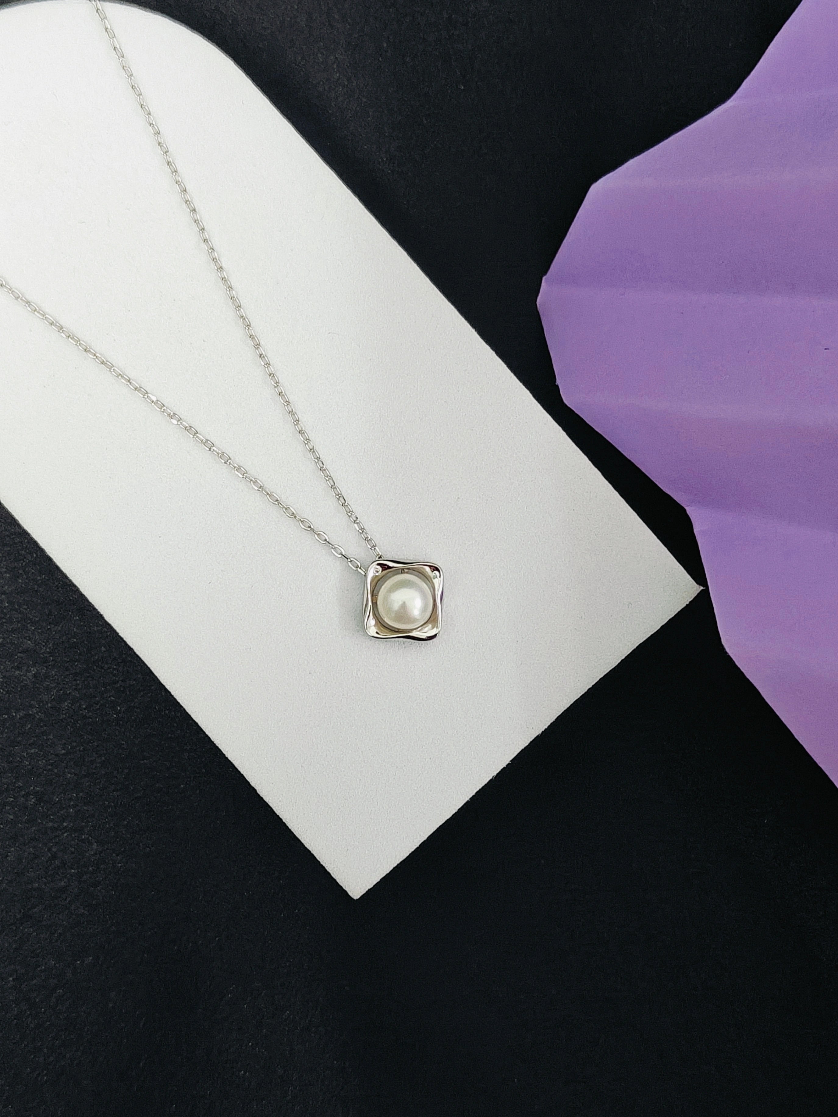 Necklace with a square pendant featuring a pearl on a white card against a black and purple background