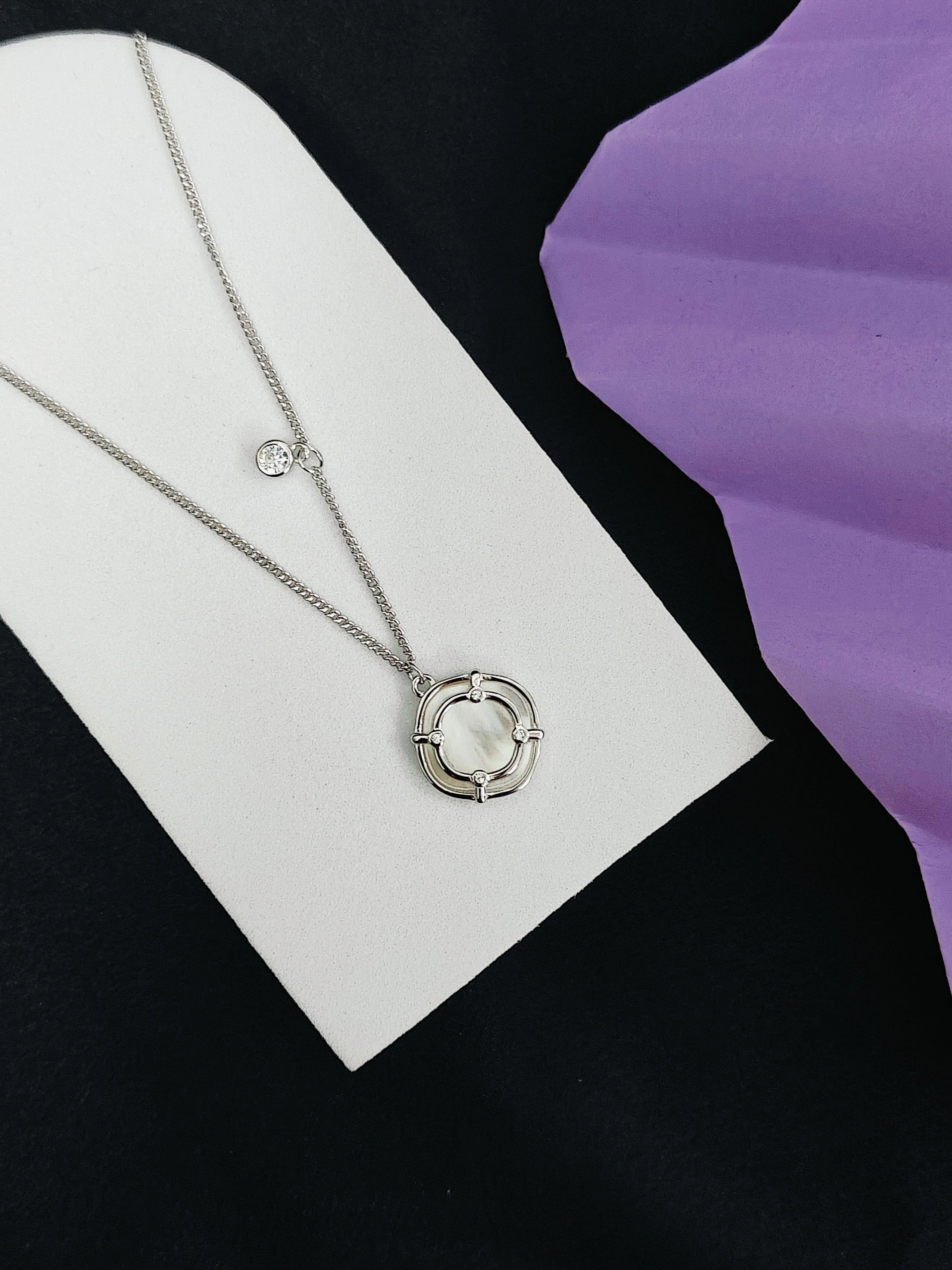 Silver necklace with a circular pendant on a white card against a black and purple background