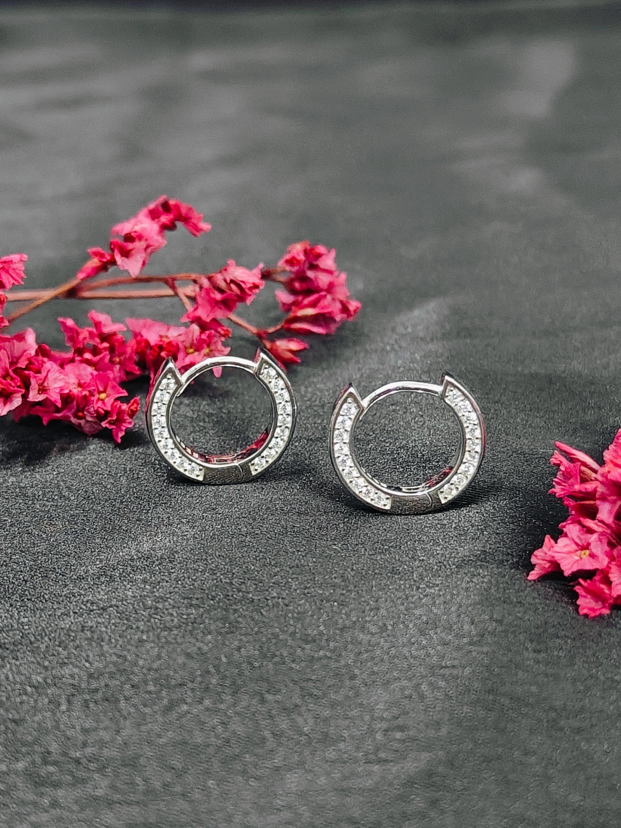 Silver hoop earrings with embedded stones on a dark surface with pink flowers.