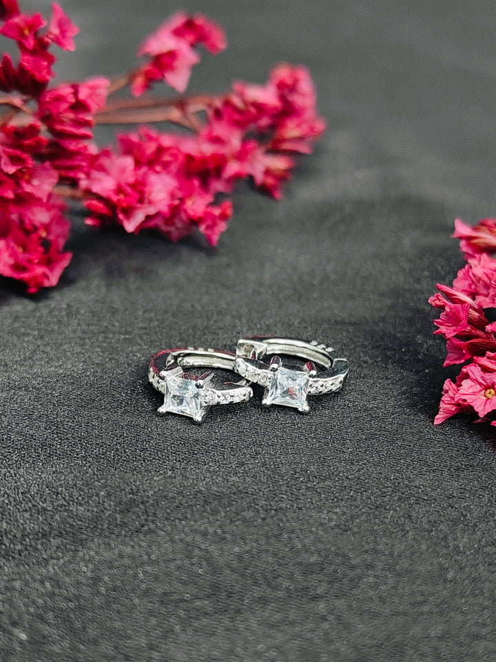 Silver hoop earrings with clear gemstones on a dark surface with pink flowers.