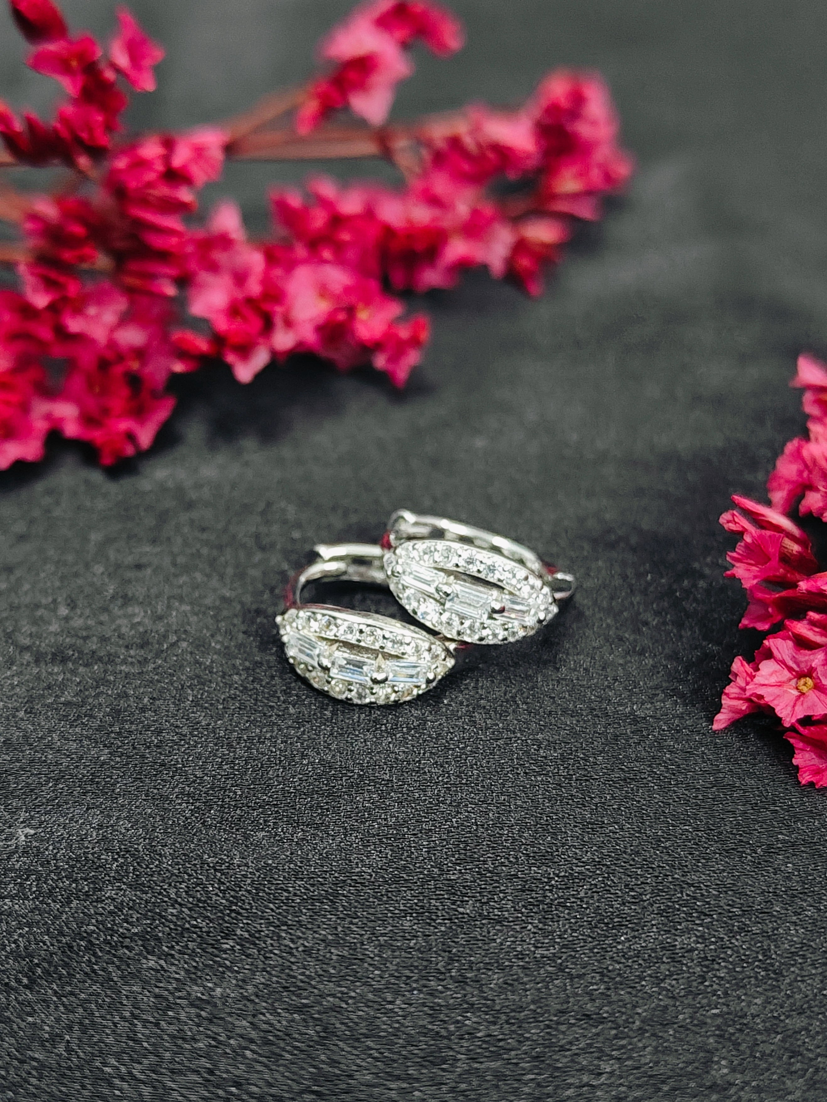 Silver wing-shaped earrings with clear stones on a dark surface with pink flowers.