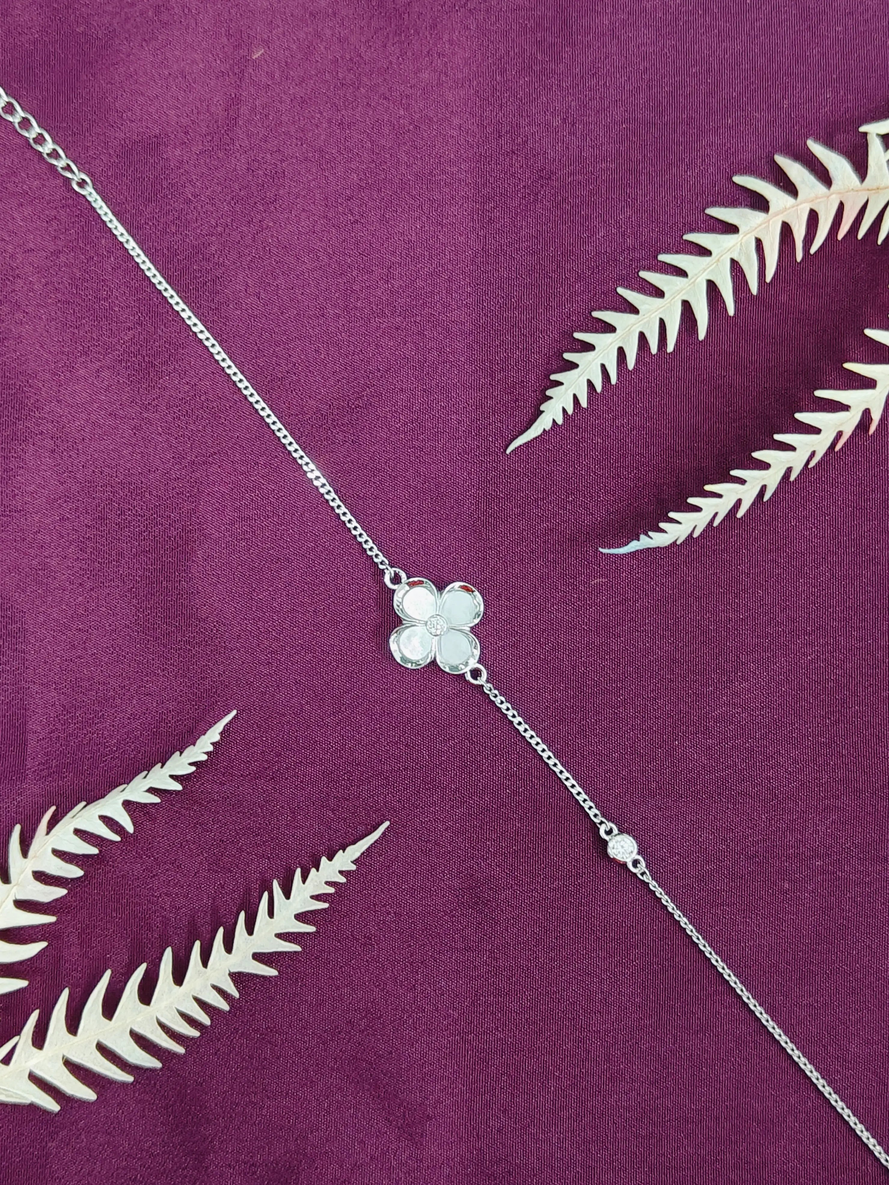 Silver necklace with a clover-shaped pendant on a purple fabric background with white leaves.