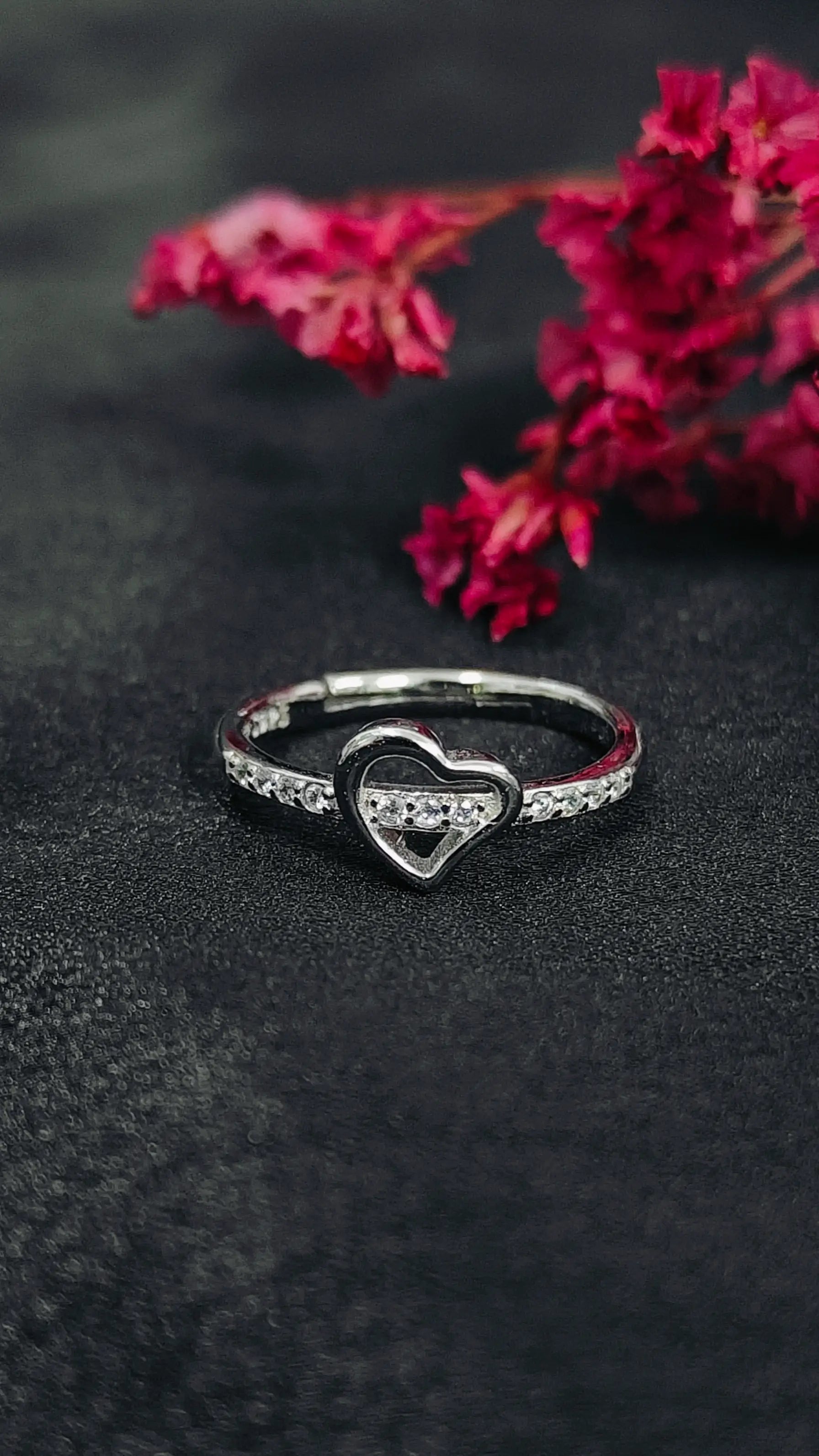 Heart-shaped ring with sparkling stones on a dark surface with pink flowers in the background