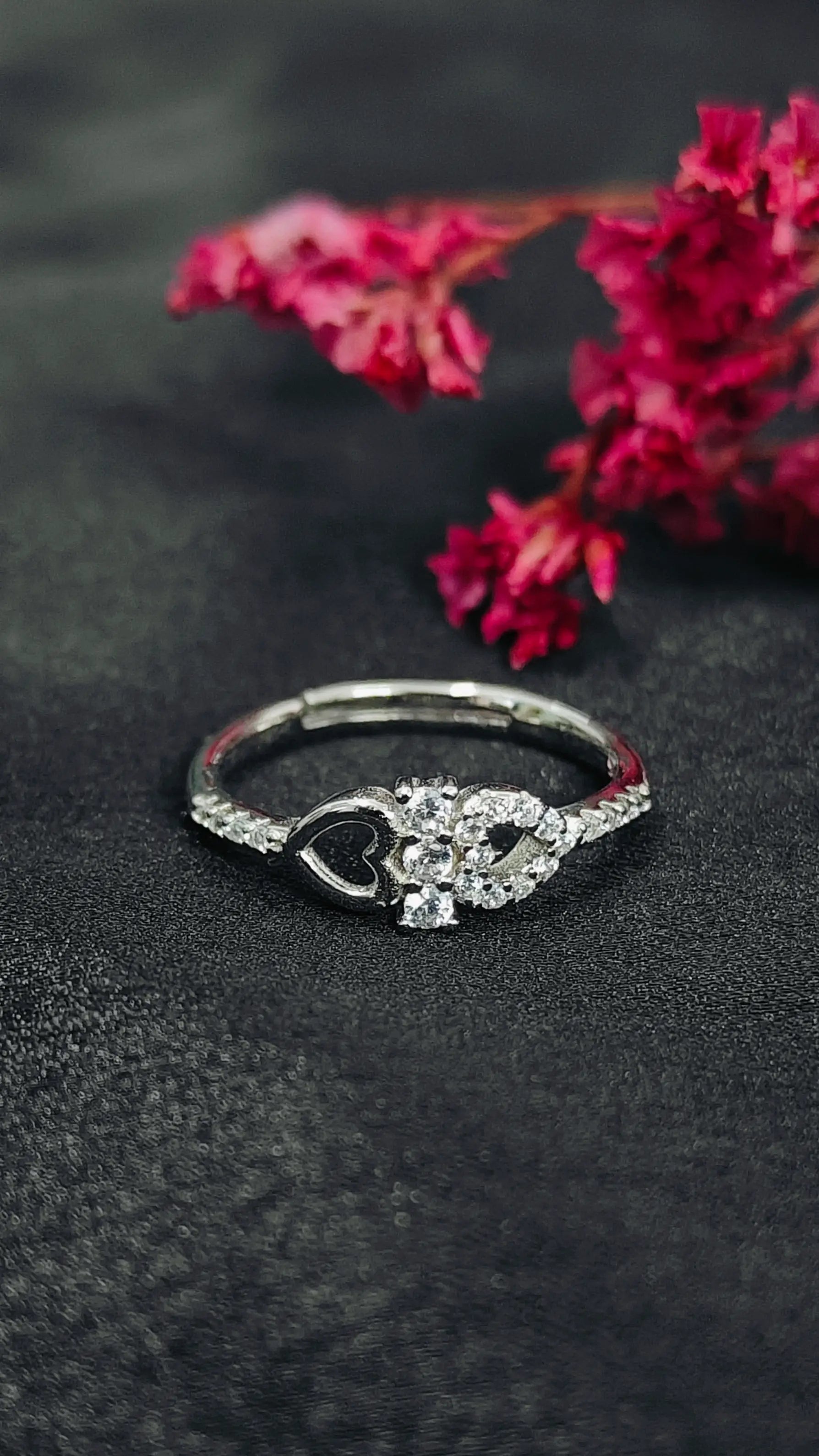 Silver ring with heart and diamond design on a dark surface with pink flowers.