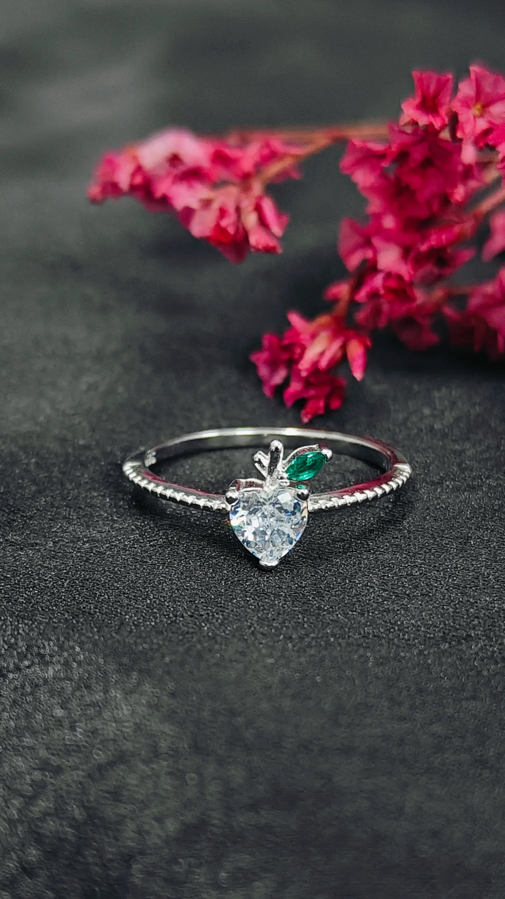 Silver ring with heart-shaped gemstone on a dark surface with pink flowers