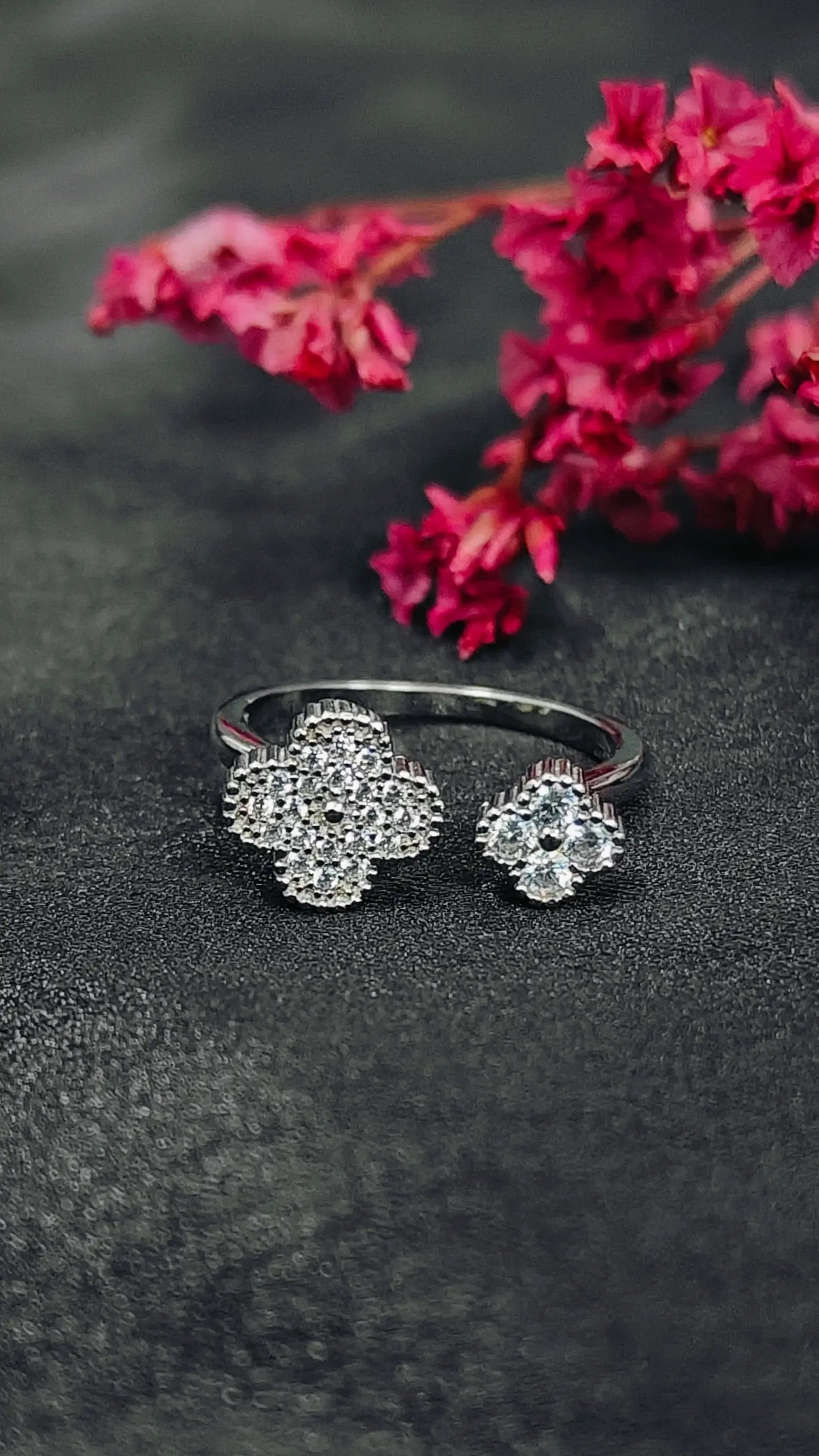 Silver ring with floral design on a dark surface with pink flowers in the background