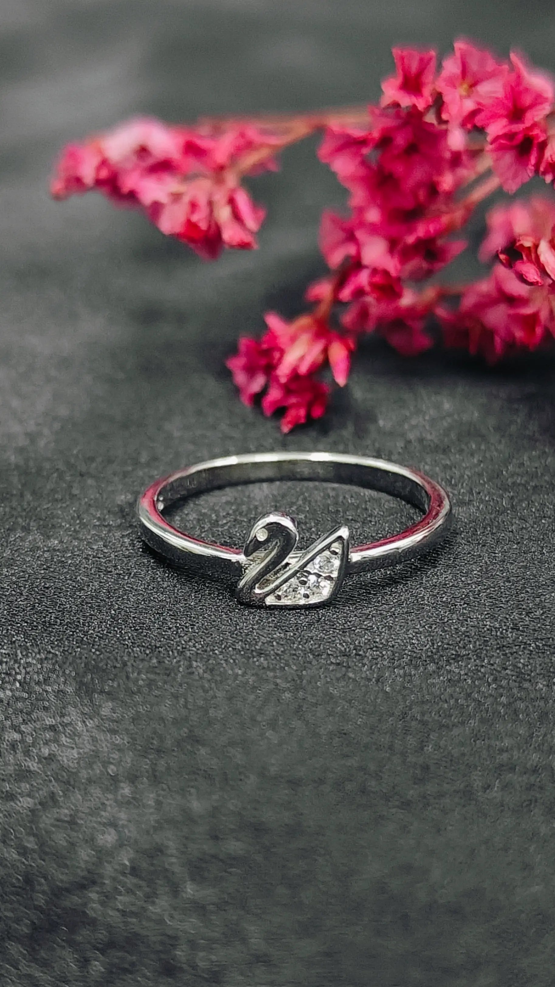 Silver swan ring on a dark surface with pink flowers in the background