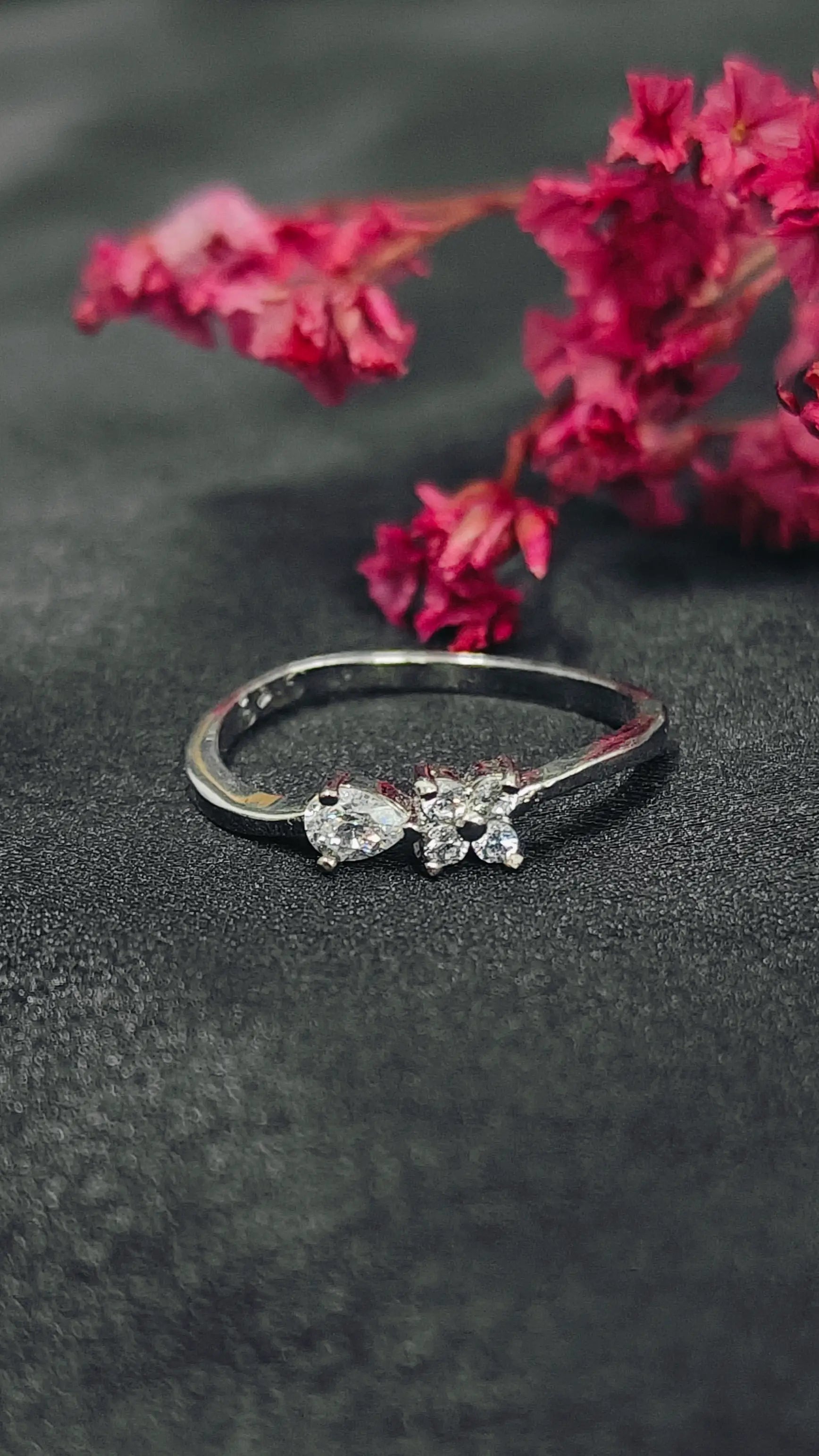 Silver ring with heart-shaped gemstones on a dark surface with pink flowers.