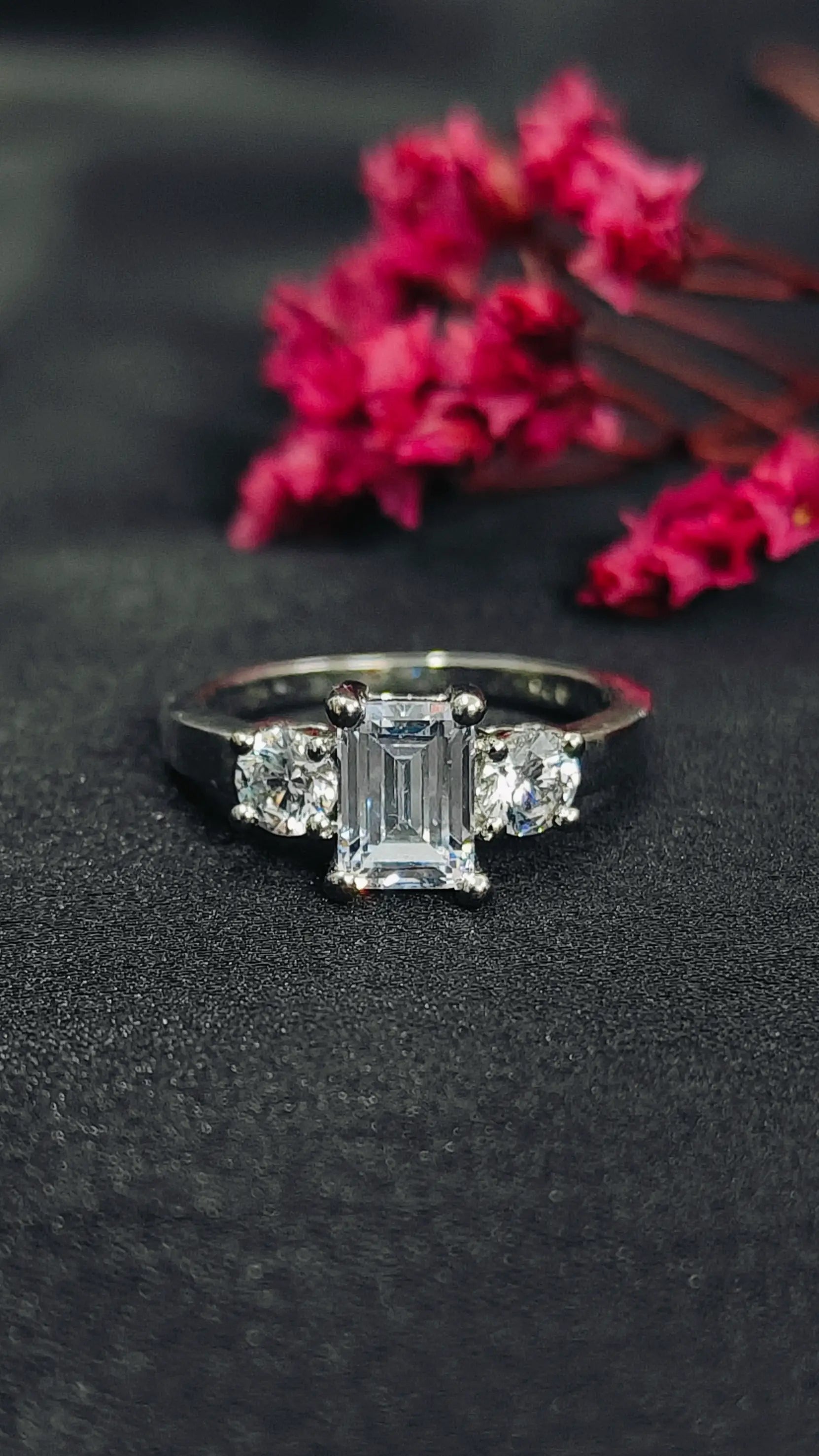 Diamond ring with emerald-cut center stone on a dark surface with pink flowers.