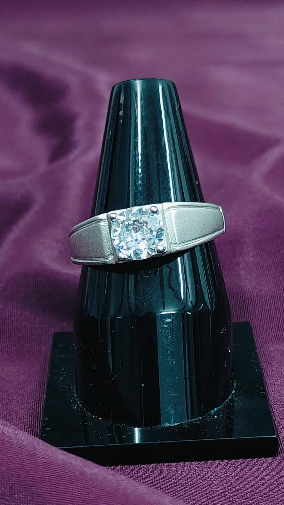 Silver ring with a diamond on a black stand against a purple fabric background